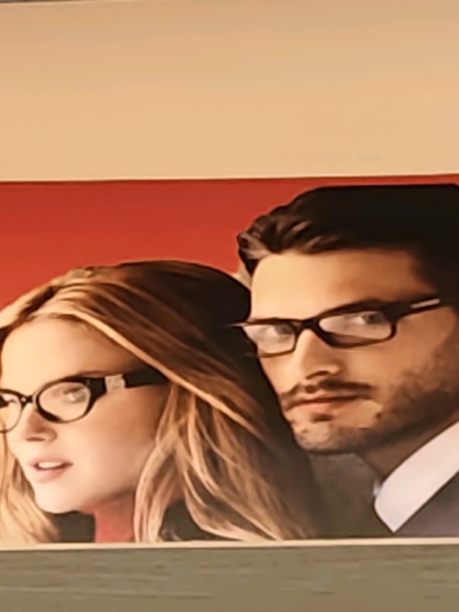 An advertisement for eyeglasses features a close-up of a man and a woman. The man, on the right, has dark hair and is wearing dark-rimmed glasses. He has a mustache and a goatee. The woman, on the left, has blonde hair and is wearing black-rimmed glasses with a decorative pattern on the temples. She is looking to her left. They are positioned against a red background. The bottom of the image shows a textured surface, possibly a wall or a display stand. The location is specified as Modhera Char Rasta, India.