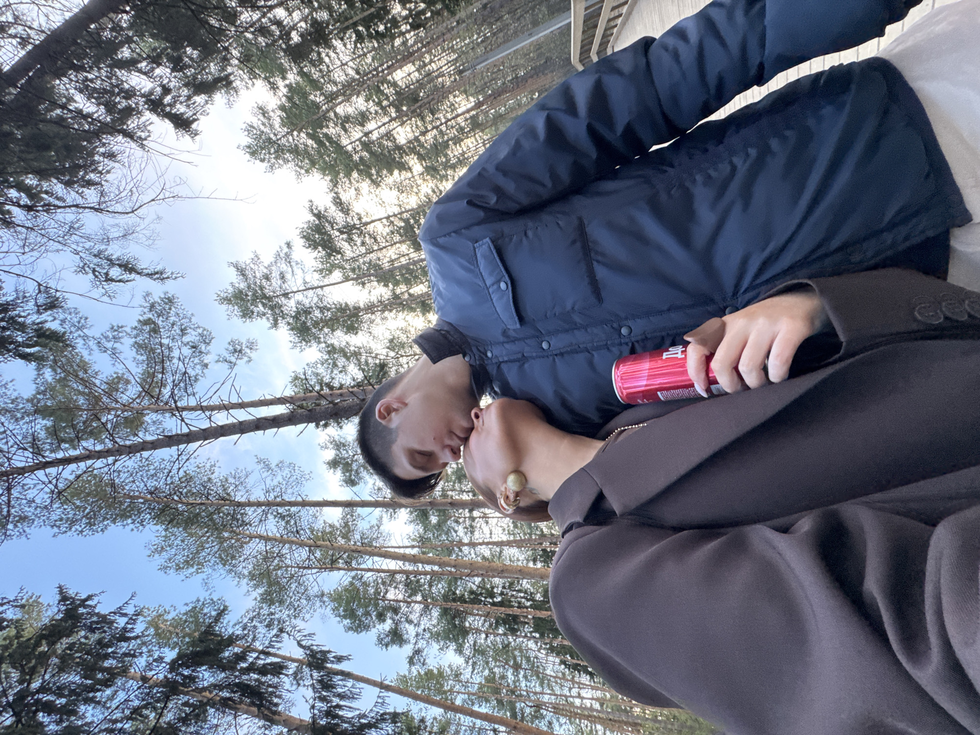 In an outdoor location near Votkinsk, Russia, a man and a woman embrace and appear to be kissing. The man wears a navy blue puffer jacket. The woman wears a brown blazer and a gold earring. The man holds a red can, possibly a beverage. Tall pine trees form a backdrop, reaching into a sky with light clouds. A wooden structure is partially visible in the background, possibly a bridge or deck.