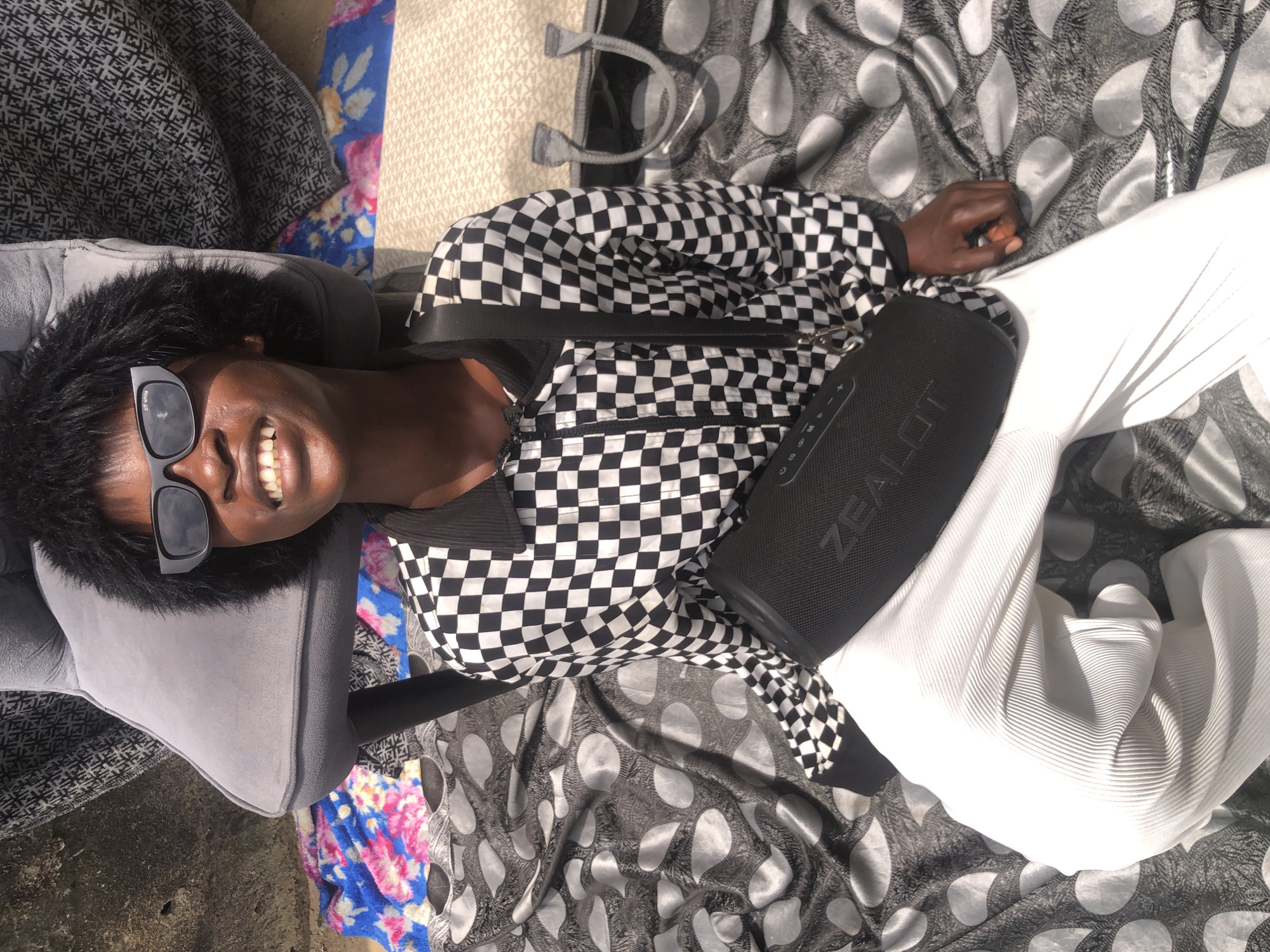 A young Black person is sitting on a patterned cloth spread on the ground, possibly in a street or outdoor area in Ado, Nigeria. They are wearing white, pleated pants, a black and white checkered jacket with a zippered front, and dark sunglasses with "off white" printed on them. A black portable speaker is positioned in front of them, and a black strap crosses their chest. A grey chair with dark patterned fabric is behind them, and other patterned fabrics are visible around them. The background appears to be a rough, textured wall. The person is smiling broadly, with their teeth visible. The lighting suggests it is daytime.