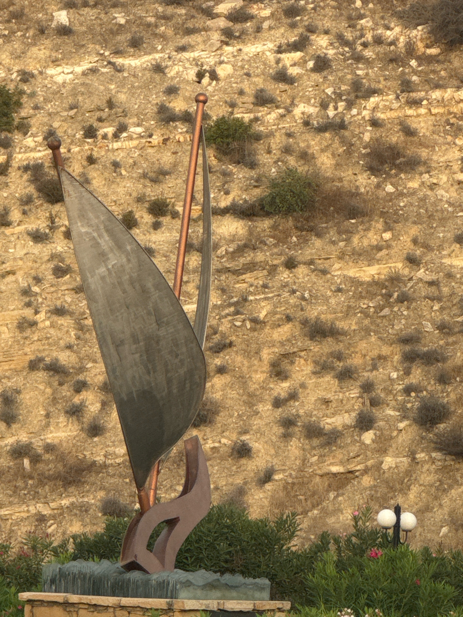 The image shows a sculpture of a sail, reminiscent of a boat, placed in front of a rocky hillside. The sculpture is made of metallic material, with a dark sail and a copper-colored mast. It is mounted on a base that appears to be a fountain with water features. In the background, the hillside is dry and sparsely vegetated with shrubs and rocks. To the right of the sculpture, a double-headed lamppost is visible, surrounded by lush green bushes and some small pink flowers. The scene is outdoors, with natural lighting suggesting it might be during the daytime. The location is identified as Agios Tychona Community, Cyprus.