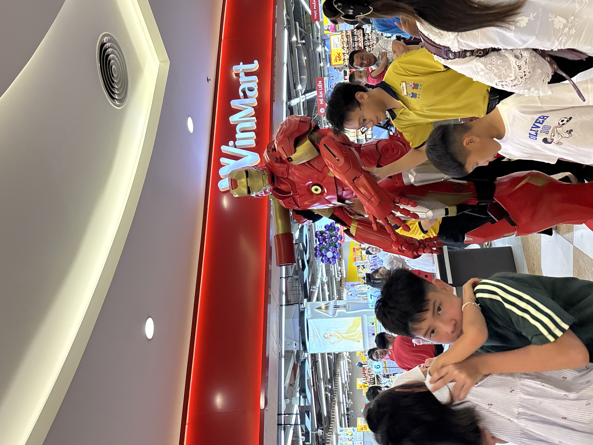 The image depicts an indoor scene within a VinMart supermarket in Đà Nẵng, Vietnam, centered around a large, vibrant red and gold Iron Man statue. Several individuals, including adults and children, are gathered around the statue, suggesting it is a public display or attraction.
In the foreground, a boy wearing a dark green t-shirt with white stripes looks towards the viewer. An adult's arm is partially visible, appearing to embrace or hold him. To the right of the Iron Man statue, another boy, dressed in a white t-shirt featuring "OLIVER 10" and a soccer player graphic, stands gazing at the figure. A person in a yellow polo shirt is also visible near the statue. The background shows various supermarket aisles with shelves stocked with products, and more shoppers can be seen.
The setting is well-lit by overhead lights, and an air vent is visible on the white ceiling. A prominent red wall on the left features the illuminated "VinMart" logo, identifying the supermarket. Visible text on signs in the background includes "THỰC PHẨM" (Food), "MÌ ĂN LIỀN" (Instant noodles), and "SÓC GIÁ" (Shock price/Sale price), indicating different product sections or promotions. Another sign reads "Saigon" with a depiction of a woman. The atmosphere suggests a typical daytime shopping environment with an added element of entertainment or novelty.