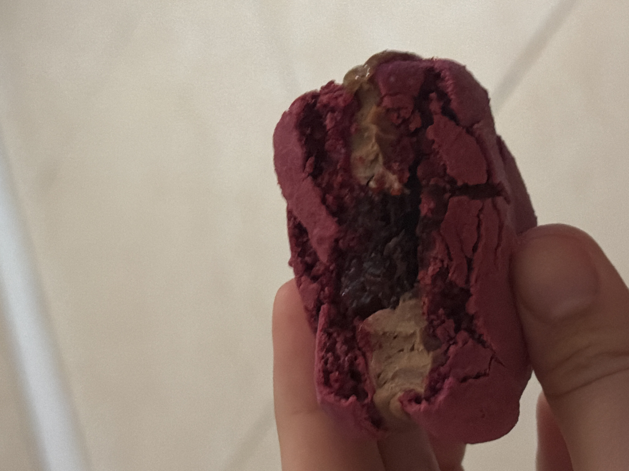 A hand holds a partially eaten, purple-colored macaron. The macaron has a light brown filling and a dark, possibly jam-like center. The background is a light beige wall. The location is not determinable from the image and is not present in the image.
