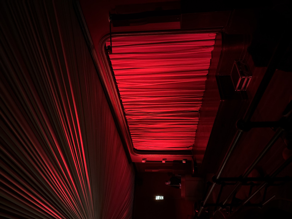 A low-angle shot bathed in red light captures a dramatic interior space. On the left, heavy, draped curtains create a radial pattern of light and shadow. Overhead, a rectangular opening reveals more draped fabric. The right side of the image descends into near-total darkness, where metal beams and possibly lighting equipment are just discernible. The intense red hue dominates the scene.