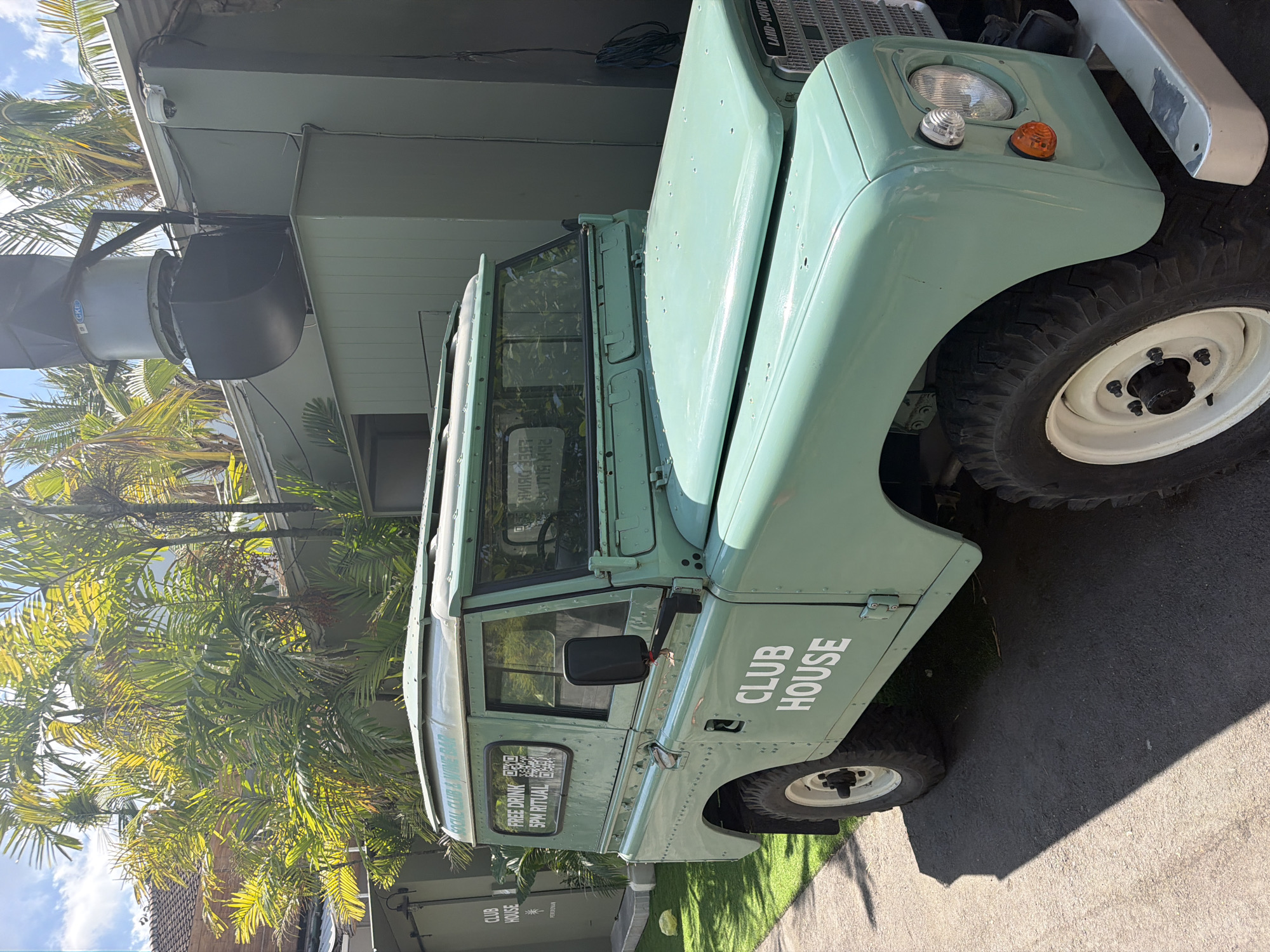 A light teal-green vintage Land Rover Defender is parked on an asphalt surface adjacent to a strip of bright green artificial turf. The vehicle features white rims on its tires, a silver bumper, and "CLUB HOUSE" emblazoned in white lettering on its side. The rear of the vehicle is not visible. Behind the Land Rover, there is a building with a light green exterior, a corrugated metal roof, and a large, industrial-looking ventilation unit on top. Palm trees with bright green fronds are visible behind and above the building, casting shadows. A small, white structure with "CLUB HOUSE" and a star symbol in white lettering is located to the left of the Land Rover, near the artificial turf. A window on the Land Rover displays text that reads "FREE DRINK 5PM RITUAL" along with a QR code. The overall scene appears to be outdoors, possibly in a cafe or recreational area, with lush vegetation suggesting a warm climate. No people are visible in the image.