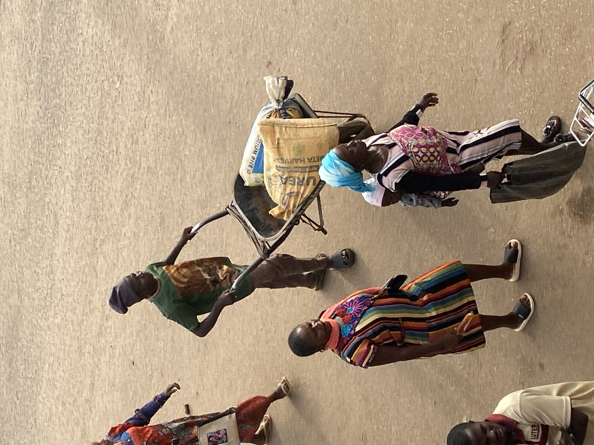 The image depicts an overhead perspective of a street or unpaved area in Bukuru, Nigeria, showing five individuals engaged in various activities. The ground is a uniform, light brown, textured surface, devoid of paved markings.
In the upper-center, a man with dark skin, wearing a green t-shirt with a brown graphic design, dark trousers, and a dark beanie, pushes a metal wheelbarrow. The wheelbarrow contains two sacks: one white sack partially visible with "MEDIUM RICE" text, and a larger yellowish-brown sack clearly labeled "UREA" and "BETA HARVEST." He wears dark sandals.
To the upper-left, partially obscured and walking away from the camera, is a person with dark skin wearing a multi-colored long-sleeved top and patterned trousers. This individual carries a light-colored tote bag with a small image printed on it and wears light-colored sandals.
In the center-left foreground, a woman with dark skin wears a vertically striped dress in multiple colors (red, orange, yellow, green, blue, black) adorned with elaborate floral embroidery in blue, pink, and orange on the chest. She has an orange scarf around her neck and holds a small, dark object in her right hand. She wears dark sandals with white soles.
To the center-right, a woman with dark skin wears a light blue headscarf and a pink and white striped dress over a long-sleeved dark undergarment. A baby, wearing a light pink hat, is secured to her back with a patterned fabric wrap. She carries a large, dark fabric bag in her left hand and wears dark sandals.
In the lower-left, a man with dark skin, wearing a light-colored polo shirt with a dark collar and the word "LUTER" visible on the chest, stands with his arms crossed, looking downwards or to the side.