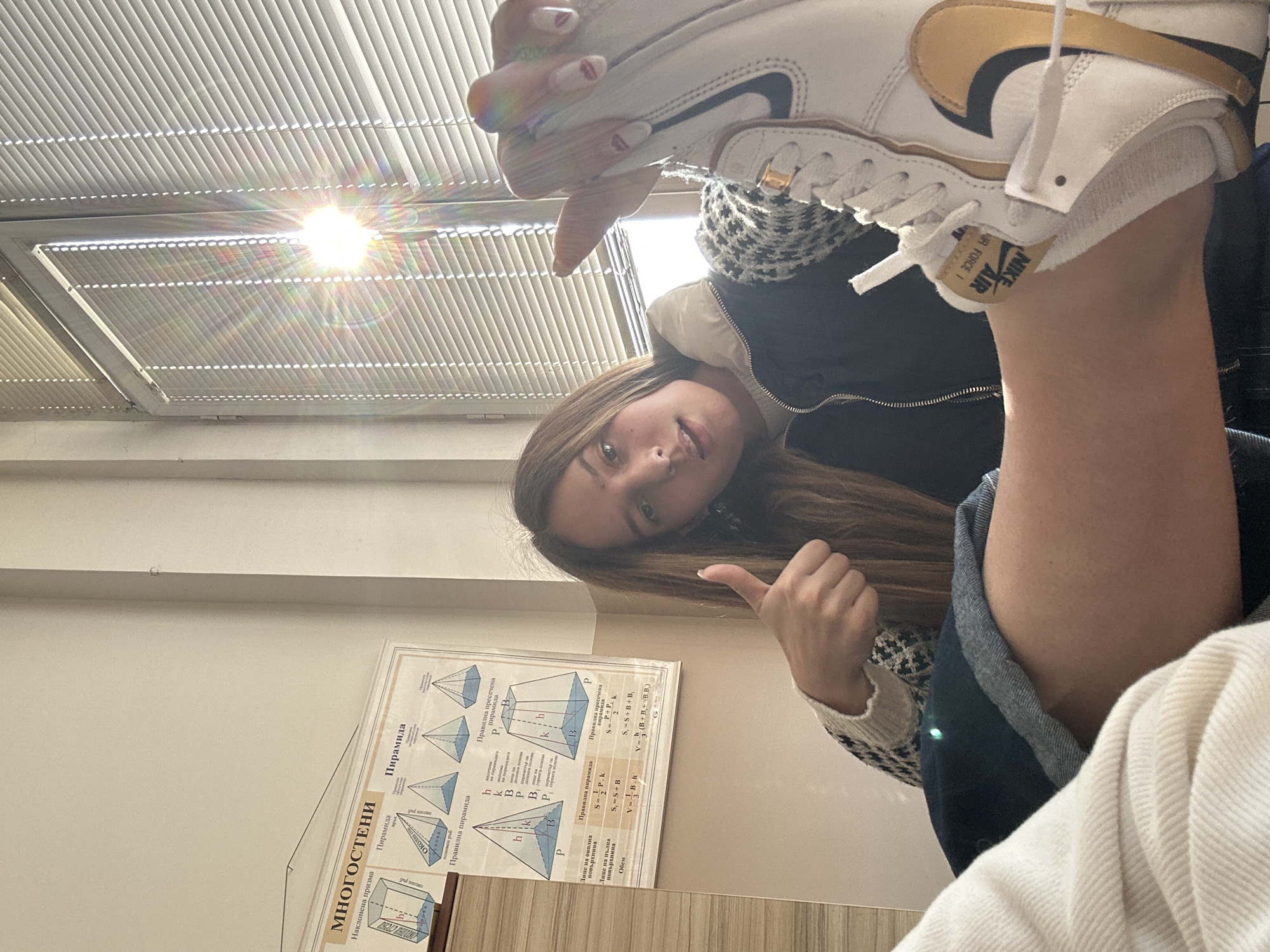 The image depicts the interior of a classroom in Sofia, Bulgaria, identifiable by Cyrillic text on a wall poster. A young female subject, with long brown hair, is positioned in the mid-ground, slightly right of center. She wears a dark blue zip-up jacket over a white and black patterned sweater. Her facial expression is neutral, with her mouth slightly ajar, as she looks directly at the camera. Her right hand is raised, forming a "thumbs-up" gesture. Her left hand is holding up a white Nike Air Force 1 high-top sneaker, which features a prominent gold-colored swoosh and a "NIKE AIR FORCE 1" label on the tongue. The fingernails on the hand holding the shoe are painted red. In the lower-left foreground, a portion of another individual's body, likely the photographer, is visible, showing a cream-colored garment and a leg clad in blue denim shorts with exposed skin. The background features a plain white wall on which a large educational poster titled "МНОГОСТЕ́НИ" (Polyhedra) is affixed, displaying various geometric shapes such as prisms and pyramids with accompanying formulas. To the right of the subject, a window with horizontal white Venetian blinds allows bright sunlight to stream into the room, creating a significant lens flare effect. The scene is brightly lit, indicative of daytime.
