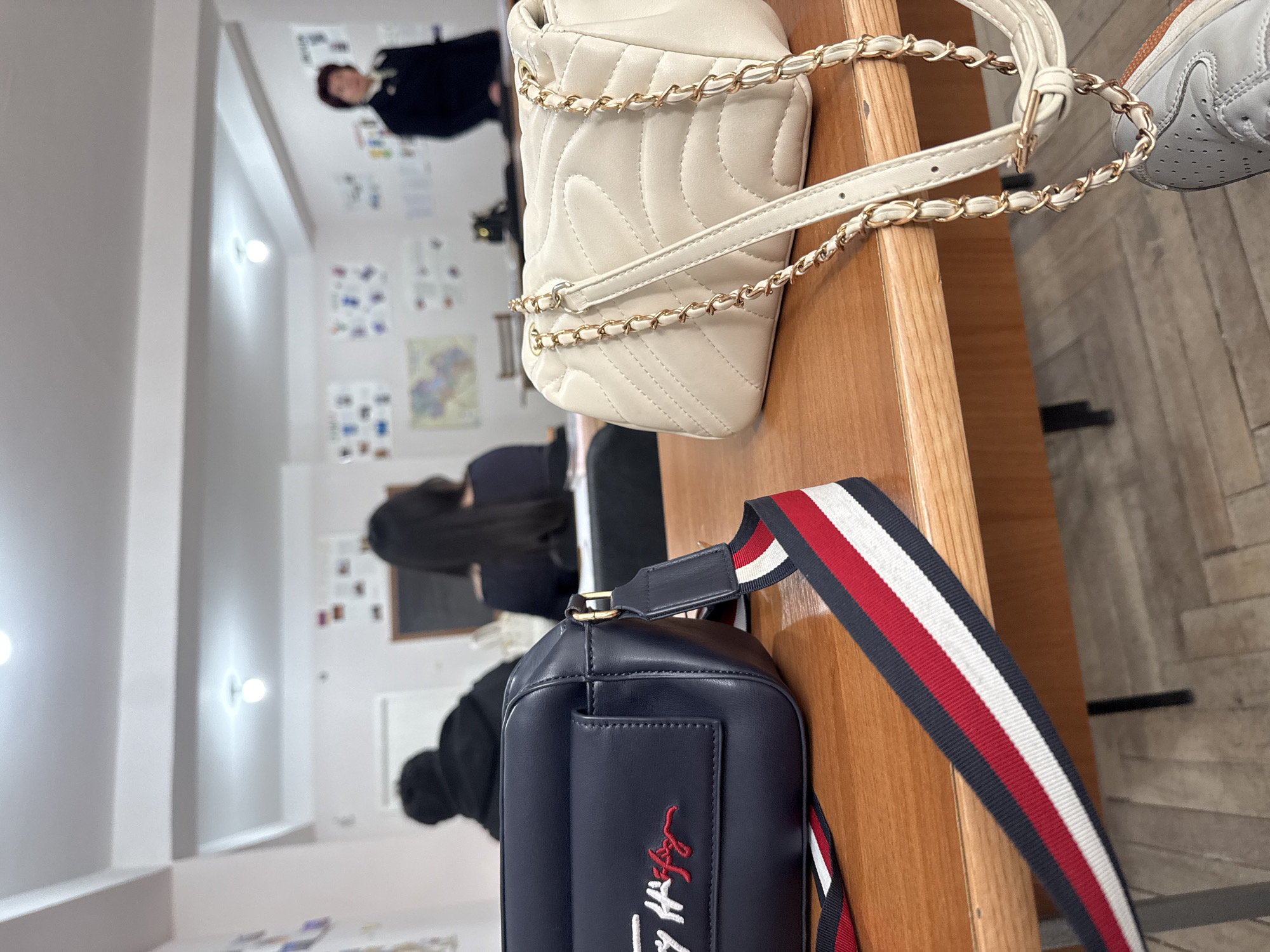 The photograph depicts a classroom or similar learning environment, most likely indoors. It shows an arrangement of objects on a wooden desk or table. In the foreground, there is a dark blue handbag with a red, white, and blue striped strap, and a white quilted handbag with a gold chain strap. A partially visible white sneaker sits near the edge of the table.
In the background, there are people seated at desks. One person has long dark hair and is facing away from the camera. Another individual is wearing a black hoodie. In the upper portion of the image, a person is standing in the background wearing a black outfit. The walls feature bulletin boards covered with various papers and materials. There is an even distribution of light in the room.
The location, Yerevan, Armenia, is not discernible from the image.