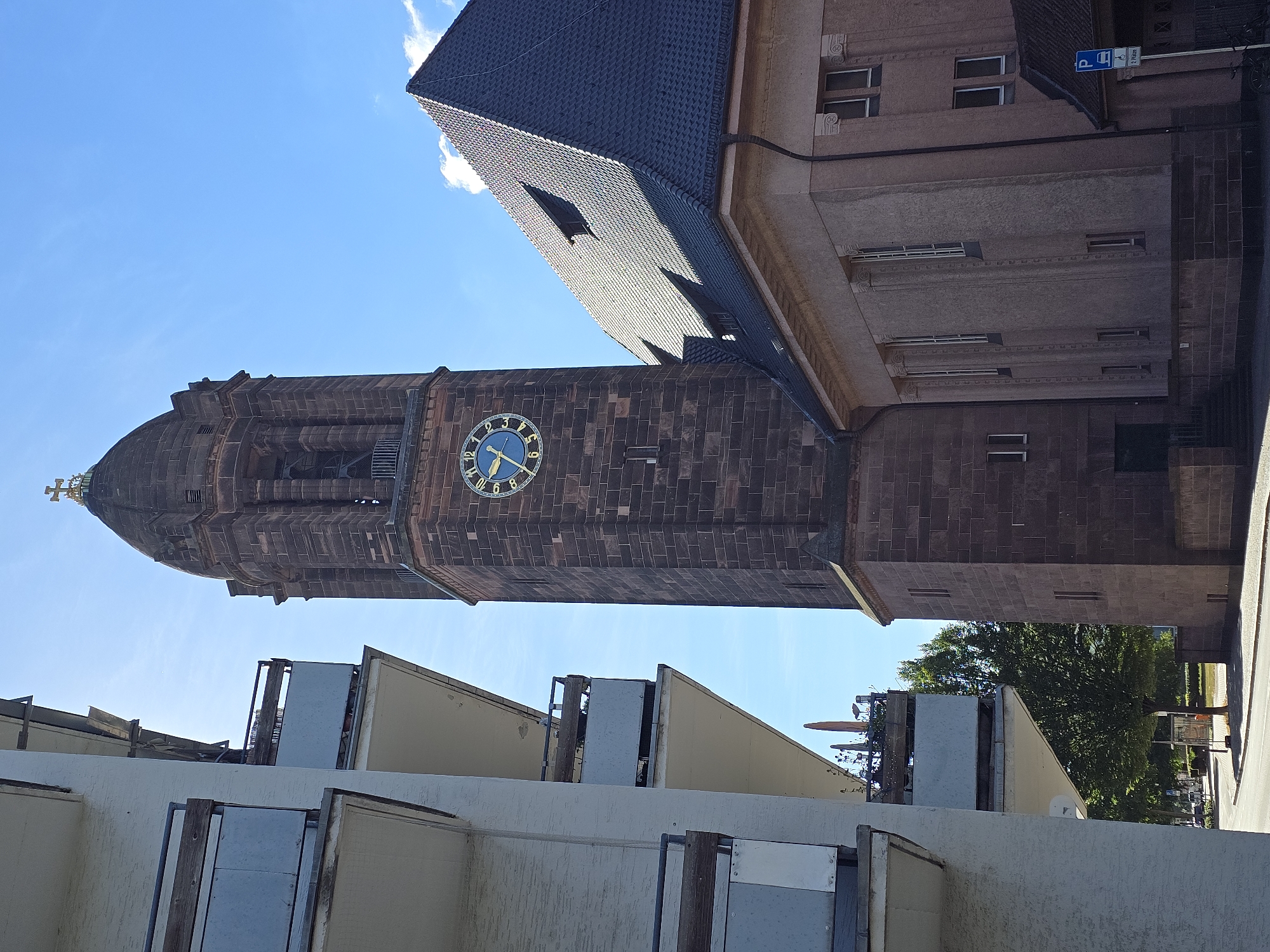 The image shows a tall, square, stone clock tower with a domed roof topped by a cross, situated next to a building with a dark, tiled roof. The clock face has Roman numerals and a blue background with gold hands. To the left of the clock tower is a modern, multi-story apartment building with balconies. The scene appears to be on a sunny day with a clear blue sky. The location is the Lutherkirche (Luther Church) in Worms, Germany, a Protestant church known for its distinctive red sandstone architecture and prominent bell tower. The building to the left is a residential building. The street in front of the church is visible, with trees and a traffic sign in the background.
