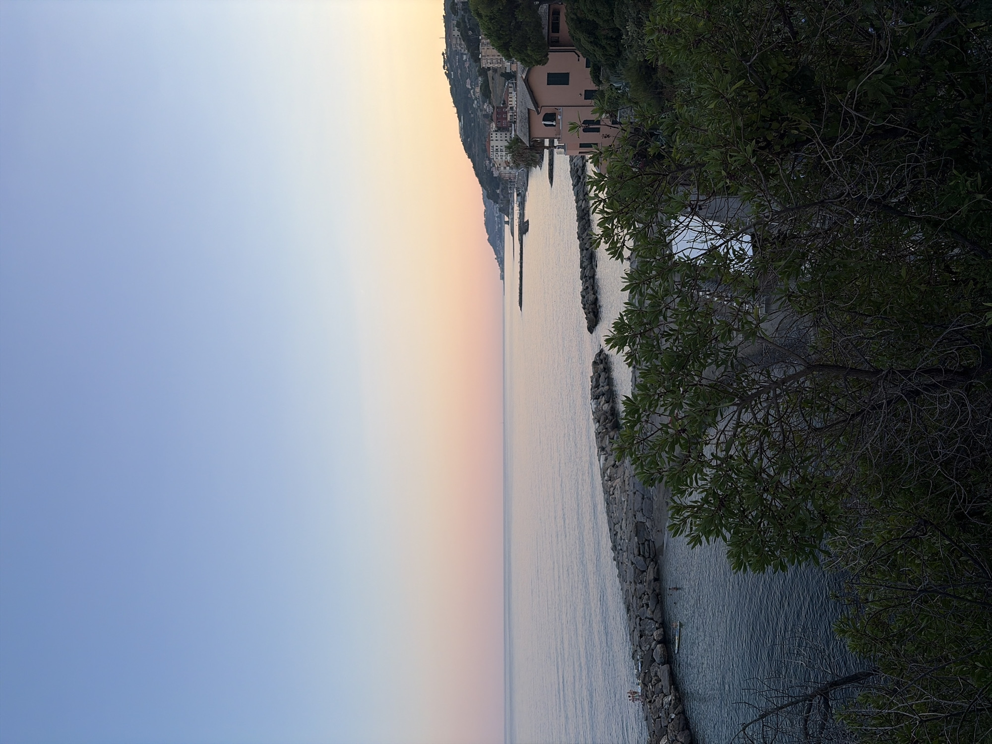 This image captures a serene coastal scene at dusk in Imperia, Italy. The sky transitions from a pale blue on the left to soft hues of pink and yellow towards the horizon, indicating the end of the day. The calm sea reflects these colors, creating a tranquil atmosphere. Along the coastline, a rocky breakwater extends into the water, providing a barrier against the gentle waves.
Dotted along the hillside in the distance, a cluster of buildings forms a picturesque Italian village, with houses painted in earthy tones. Dark green foliage, likely trees and shrubs, frames the right side of the image, adding depth and a sense of lushness to the landscape. The overall impression is one of peacefulness and natural beauty. The time of day is clearly sunset, and the weather appears clear and calm. While no specific text is visible, the architecture and the seaside setting strongly suggest a Mediterranean location.