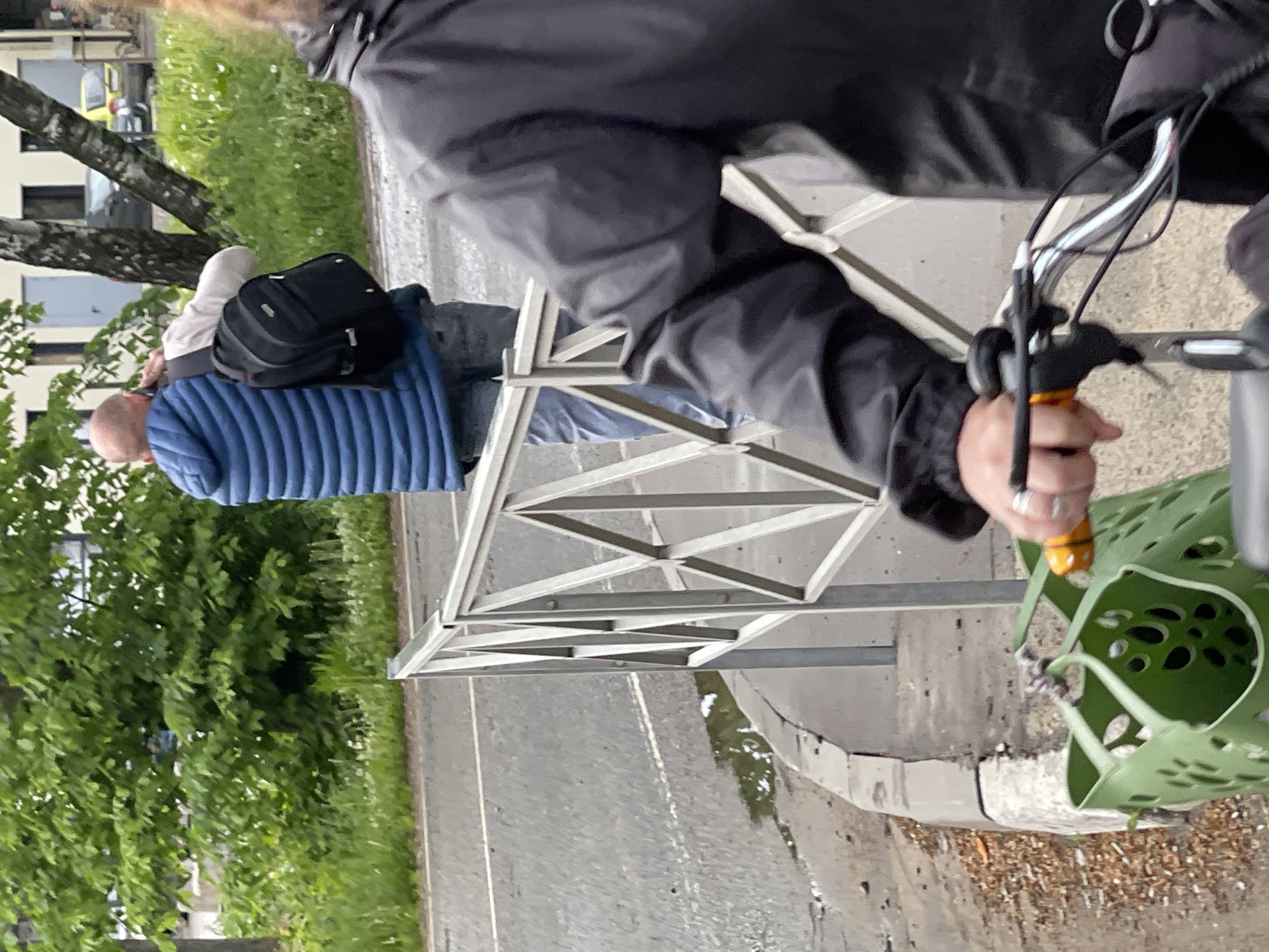 A view of a bicycle rider in Ivanovskoye, Russia shows the right hand and portion of the torso of a person wearing a black jacket on a bicycle with a green basket, standing next to a gray metal fence bordering a road. In the background, separated by the fence, is a man with a shaved head and wearing a blue puffer jacket and a black backpack. Green trees are also visible in the background, with building structures in the distance. The road appears to be wet, with puddles along the curb.