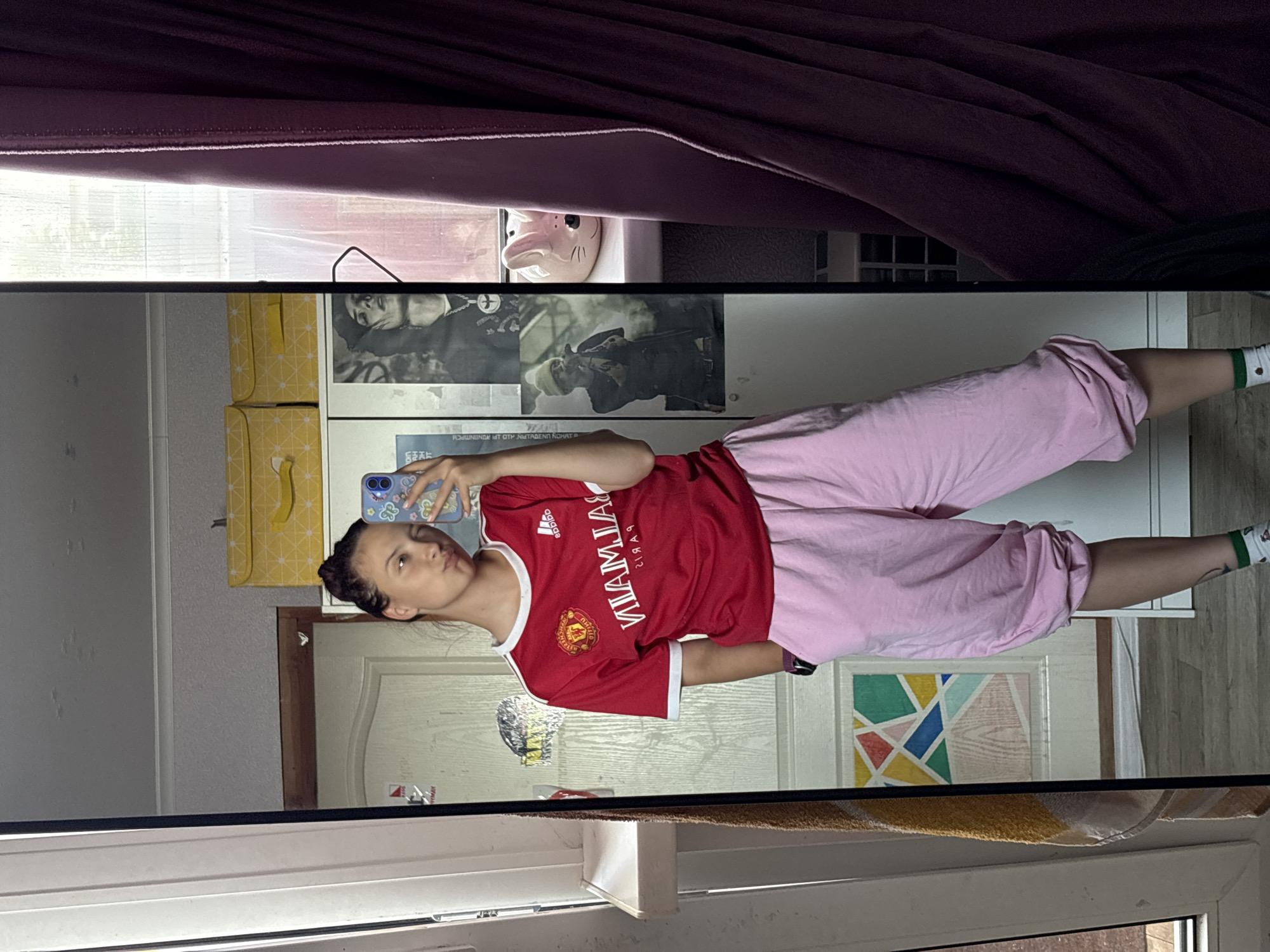 A person wearing a red Manchester United football jersey and pink sweatpants is taking a mirror selfie. The location is likely an indoor space, but its description is limited due to the mirror's reflection. A piggy bank sits atop a white cabinet. The city cannot be determined.