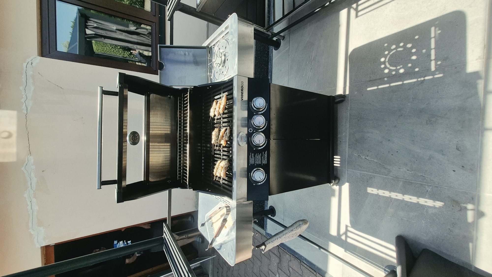 A black and silver gas grill is positioned on a grey tiled patio. The grill is open, and several skewers of food, likely sausages or meat, are cooking on the grates. A plastic container with tongs sits on the left side of the grill. In the background, a white wall with visible cracks runs across the upper portion of the image. A window with dark brown trim reflects an outdoor scene with greenery and a covered structure. To the left of the grill, a portion of a metal table and a person in a dark shirt are visible, suggesting this location is a residential outdoor area. The sunlight casts distinct shadows of the grill and railings onto the patio tiles. There is no direct information to specify the location within Chemnitz, Germany.