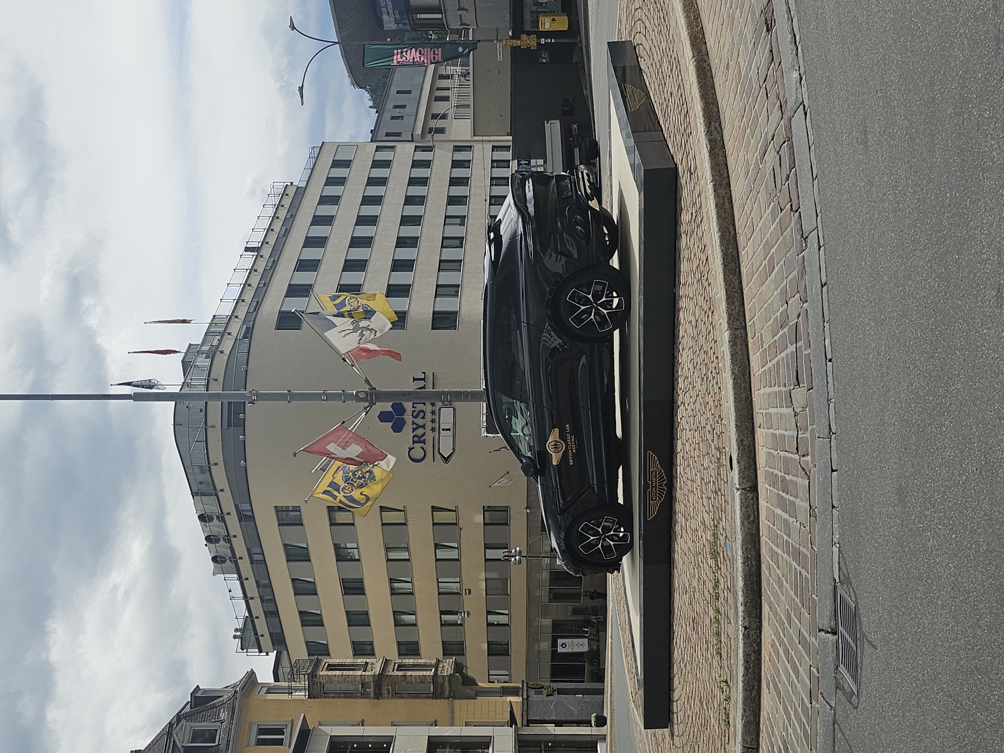 A black Aston Martin is displayed on a raised platform in front of a light-colored building. The building has multiple floors with rows of windows and a curved facade. Several flags are visible, including the Swiss flag and what appears to be a yellow and blue crest flag, as well as a red flag. The Aston Martin has a gold emblem on its side that reads "BRITISH CLASSIC CAR". A black sign with the Aston Martin logo is also visible next to the car. The ground in front of the building is paved with bricks, and a paved road runs alongside it. The sky is cloudy. The scene appears to be an outdoor display in a town or city, likely St. Moritz, Switzerland, given the contextual information. The time of day appears to be daytime, with bright lighting. No people are clearly visible in the frame.