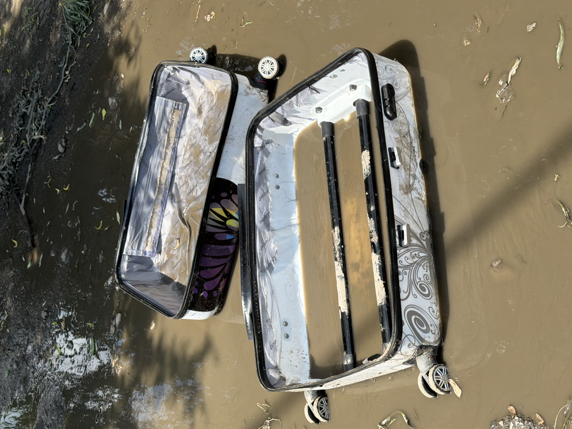 The image shows two suitcases partially submerged in muddy water, likely floodwater. One suitcase is white with a swirling pattern, and the other is decorated with a purple and black butterfly design. Both suitcases are open and appear to be damaged. The setting appears to be outdoors. The location of Dinkata, Bulgaria, cannot be confirmed from the image. No people are visible in the image.