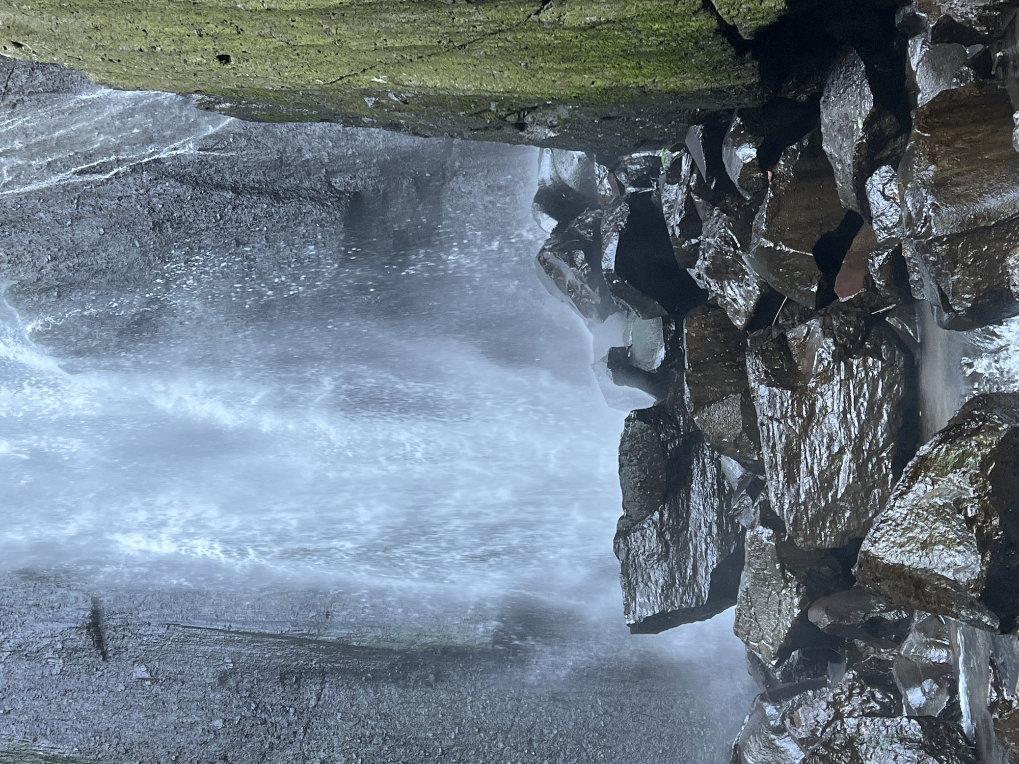 The image shows a waterfall in Busungbiu, Bali, Indonesia. The waterfall cascades down a rocky cliff face, with water spraying and misting around the rocks at the base. The rocks are dark and wet, with some appearing to be covered in moss on the right side of the image. The water creates a shimmering effect as it falls, and the overall scene conveys a sense of natural beauty and power.