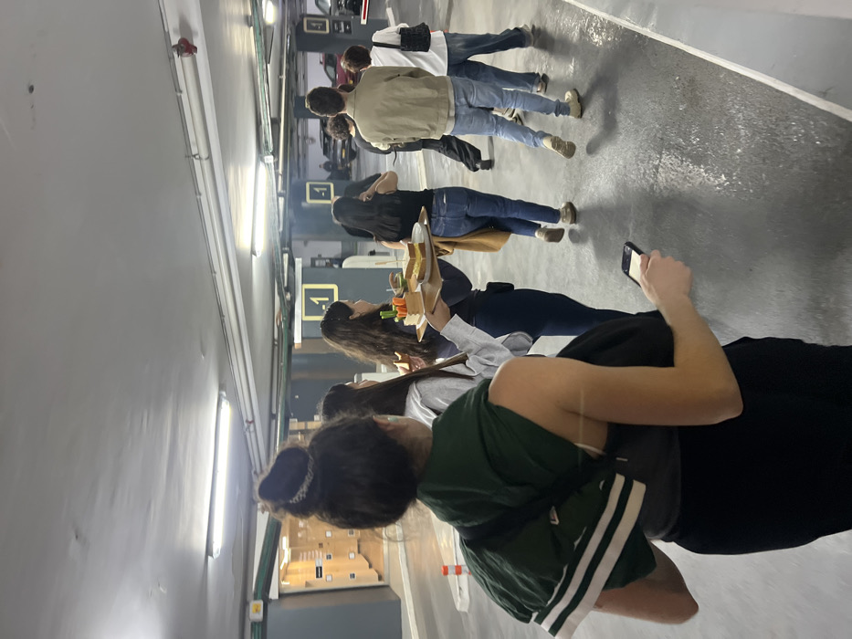The image depicts a group of approximately eight individuals walking through a brightly lit underground parking garage, identified as level -1, in Tel-Aviv, Israel. The environment features a grey concrete floor with white painted lines, and a concrete ceiling with exposed pipes, electrical conduits, and fluorescent strip lighting.
In the foreground, a female with dark hair tied in a bun, wearing a dark top and black pants with a dark green jacket draped over her left shoulder and a cross-body bag, holds a black smartphone in her right hand. Immediately to her right and slightly ahead, another female with long dark hair carries a wooden tray containing two large layered sandwiches, a small wooden container filled with cut vegetables (carrots and celery), and a glass containing a beverage.
Further into the frame, a cluster of approximately six additional individuals, appearing to be young adults, walk in the same general direction away from the camera. They are dressed in casual attire, including jeans and light jackets. One male in the mid-ground wears a light brown jacket and blue jeans. In the background to the left, a series of light-colored modular storage units or lockers are visible along a wall. To the right in the background, parked vehicles are partially discernible. Two orange traffic cones are positioned near a wall on the left side of the frame. The group appears to be in transit through this utilitarian space.