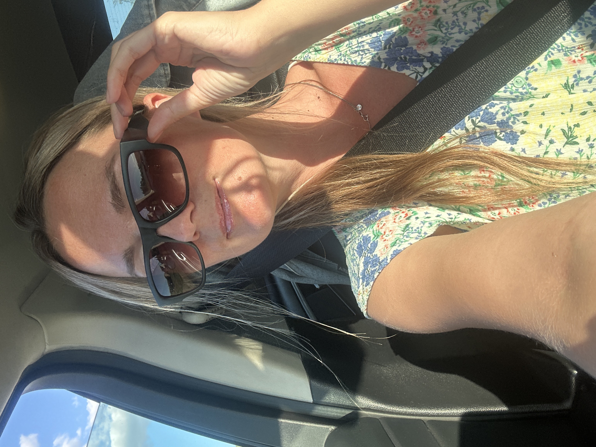 A blonde woman wearing sunglasses and a floral print dress is sitting in the passenger seat of a car. She is adjusting her sunglasses with her right hand. Her hair is long and blonde, and it is partially obscuring her face. The car appears to be in motion, as indicated by the blurred background. The sky is blue with some clouds. The woman is wearing a seatbelt. The image appears to have been taken during the daytime. The location is Novotolucheyevo, Russia.