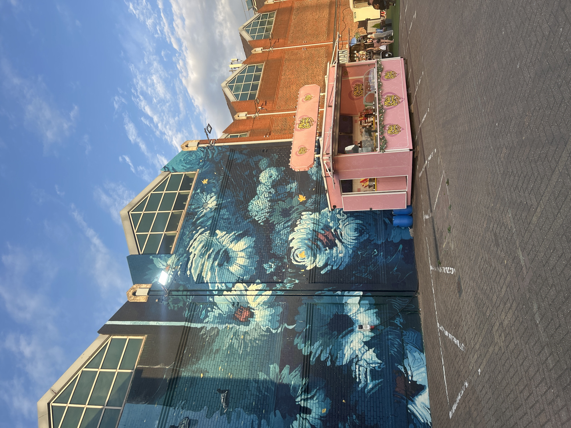 The image depicts a street scene in London, United Kingdom. A large building with a floral mural covers its wall. Next to the building is a pink "Miss Churros" food stand. The foreground shows a paved parking area with white parking lines. In the background, there is a red brick building with uniquely shaped windows and a small group of people in a grassy area. The sky above shows a blue sky with scattered clouds.