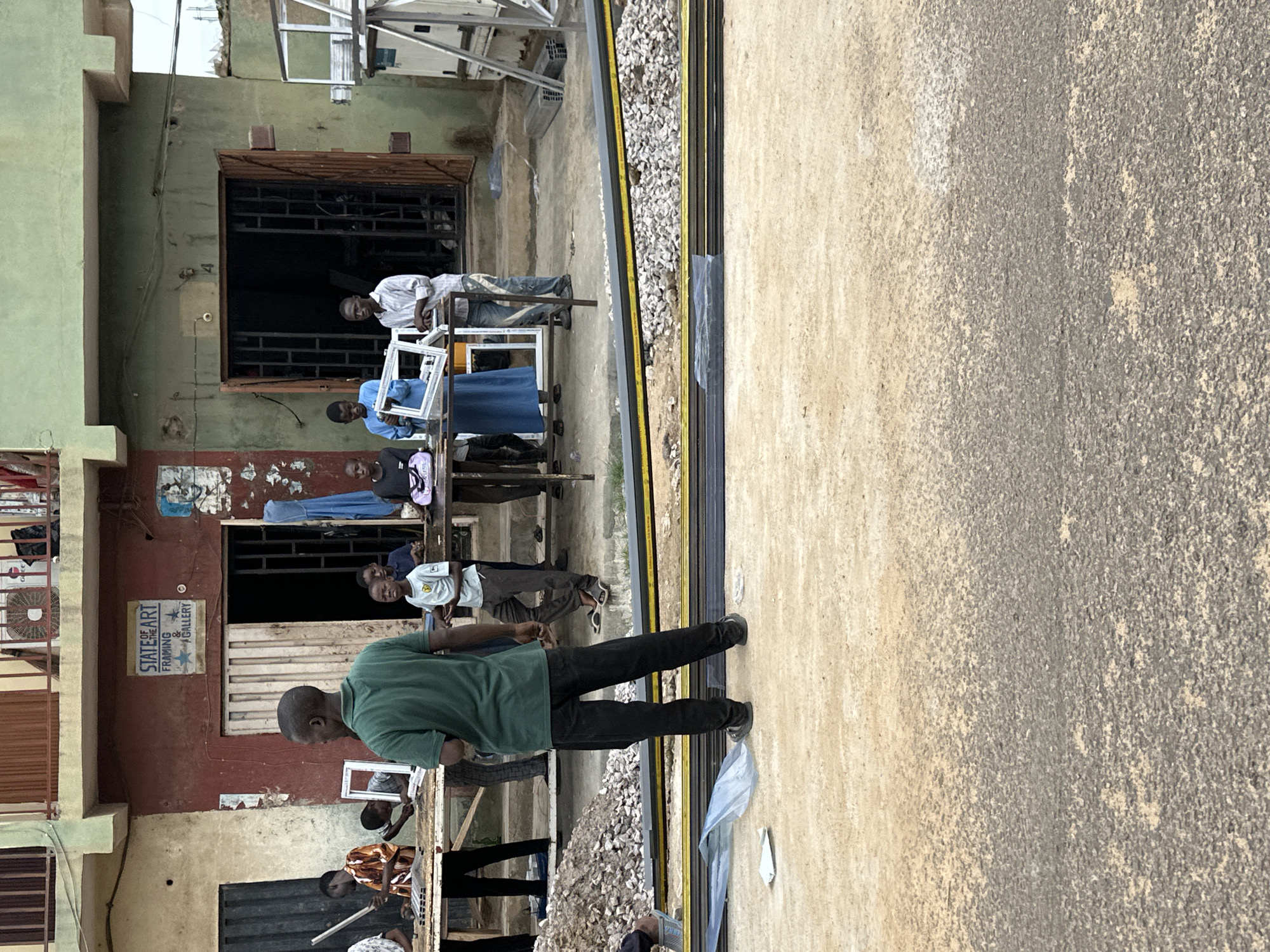 The image depicts a street scene outside a shop in Kaduna, Nigeria. Several individuals are present, appearing to be engaged in work or business. In the foreground, a pile of gravel and what appears to be building materials, possibly aluminum profiles, are stacked on the ground. A few people stand around tables laden with frames and other items.
A man in a green shirt and black pants is facing away from the camera, observing the activity. To his left, another man in an orange and white patterned shirt is holding a long object, possibly a tool. Further left, a young boy is looking at a framed object on the table.
In the center of the image, a group of people, including men and boys, are gathered around tables. One man is holding a white frame. Another is wearing a blue dress or robe.
To the right, a man in a striped shirt and jeans stands near an open doorway. In the background, the building appears to be a shop with a sign that reads "STATE OF THE ART FRAMING & GALLERY." The shop has metal grilles on its windows and doors. The overall environment suggests an outdoor workspace or marketplace.