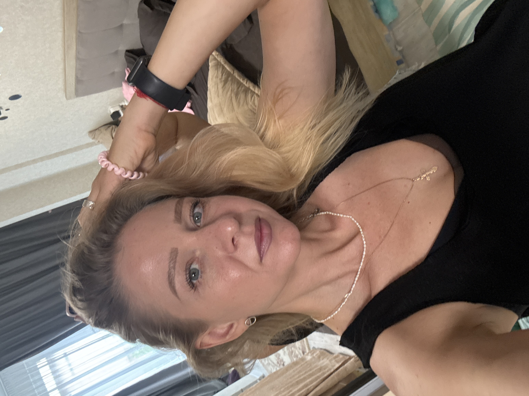 A woman with blonde hair and blue eyes is taking a selfie in a bedroom. She is wearing a black tank top, a pearl necklace, and a cross necklace. She has on a smartwatch on her left wrist and a pink spiral bracelet on her right wrist. Her right hand is raised, touching her hair. The background shows a bed, curtains, and a dresser.