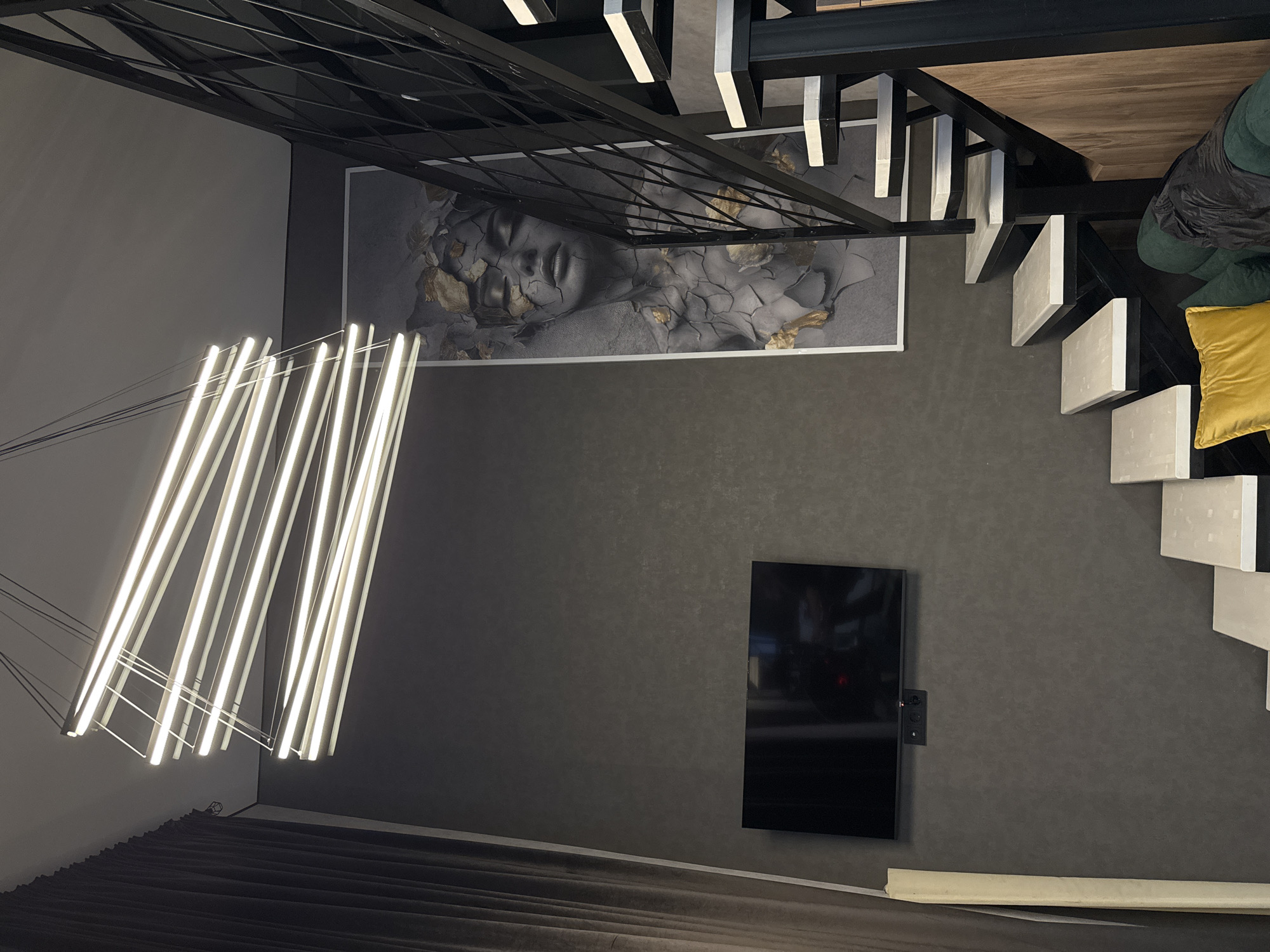 The image depicts an interior space with a mix of modern and artistic elements. Dominating the upper portion of the frame is a black metal structure, possibly part of the building's architecture or a decorative fixture. Adjacent to this is a large piece of artwork featuring a grayscale image of a face with cracks filled with golden accents, adding a touch of elegance.
To the right of the artwork, a staircase with white steps and a black metal railing ascends, partially covered by dark green pillows and a bright yellow cushion, suggesting a seating area. Below the artwork, a flat-screen television is mounted on the wall, providing a modern entertainment feature.
In the lower left corner, a set of linear LED lights hang from the ceiling, illuminating a dark gray curtain or fabric, indicating a window treatment or room divider. A strip of white trim runs along the bottom of the wall, adding a subtle contrast to the gray walls and flooring. The overall color palette is subdued, with grays, blacks, and whites creating a sophisticated atmosphere, punctuated by the gold accents and the bright yellow cushion. The location is unknown. There are no people, actions, or interactions visible in the image.