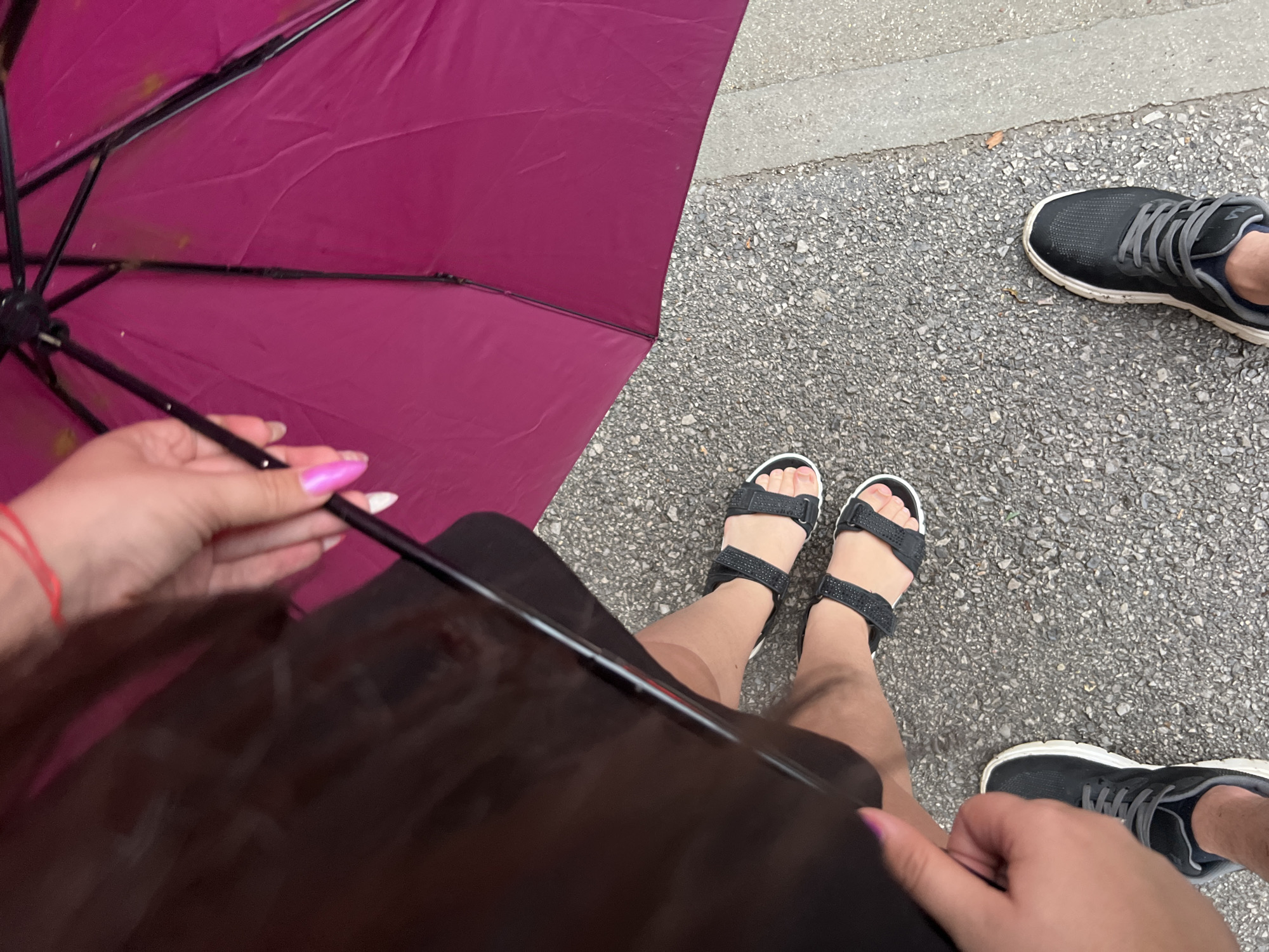 This is an overhead shot of two people's legs and feet. One person is wearing black sandals and the other is wearing black sneakers. The person in the sandals is closer to the camera and is holding a dark object, possibly a tablet, in their lap. A portion of a magenta umbrella is visible in the upper left corner of the frame. The ground appears to be asphalt or pavement with scattered pebbles. The scene is outdoors, with the paved ground suggesting a street or walkway. The lighting indicates it is daytime. No visible text is present in the image. The overall impression is a casual outdoor scene, perhaps a break from walking or an outdoor gathering.