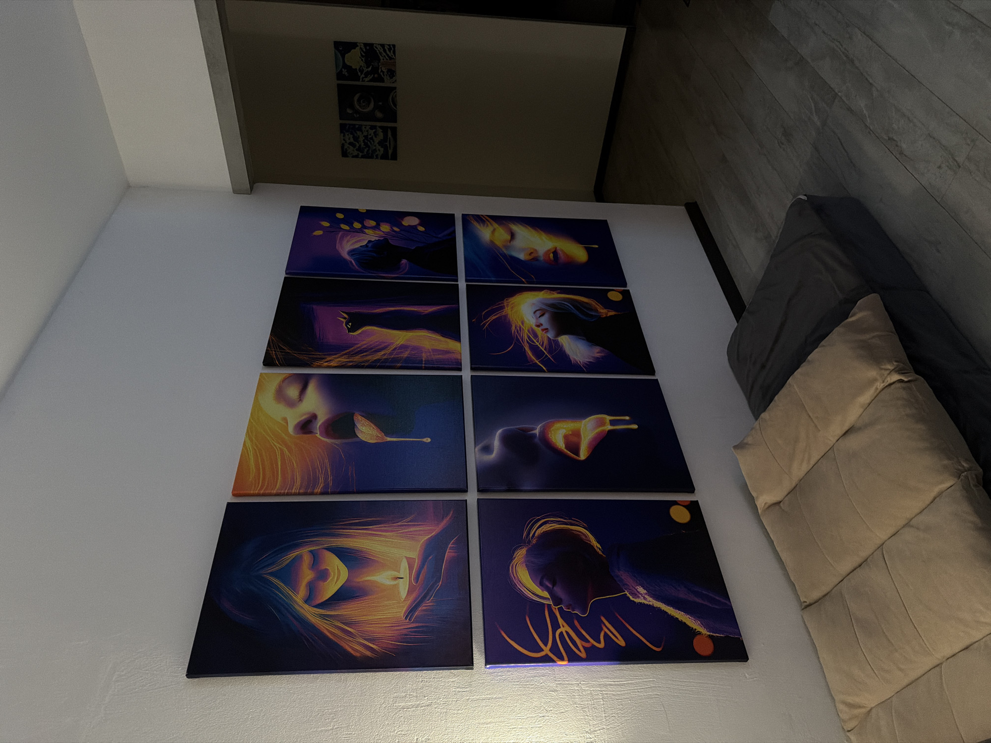 The image shows a living room with a wall adorned with eight framed artistic prints arranged in a two-by-four grid. The prints depict stylized portraits of women with glowing, vibrant colors. In front of the wall, a floor mattress with beige and gray cushions is placed. The room has a light-colored textured wall and a dark wooden floor. A doorway in the background leads to a darker area. The city of Naberezhnye Chelny, Russia, is not mentioned or depicted in this image.