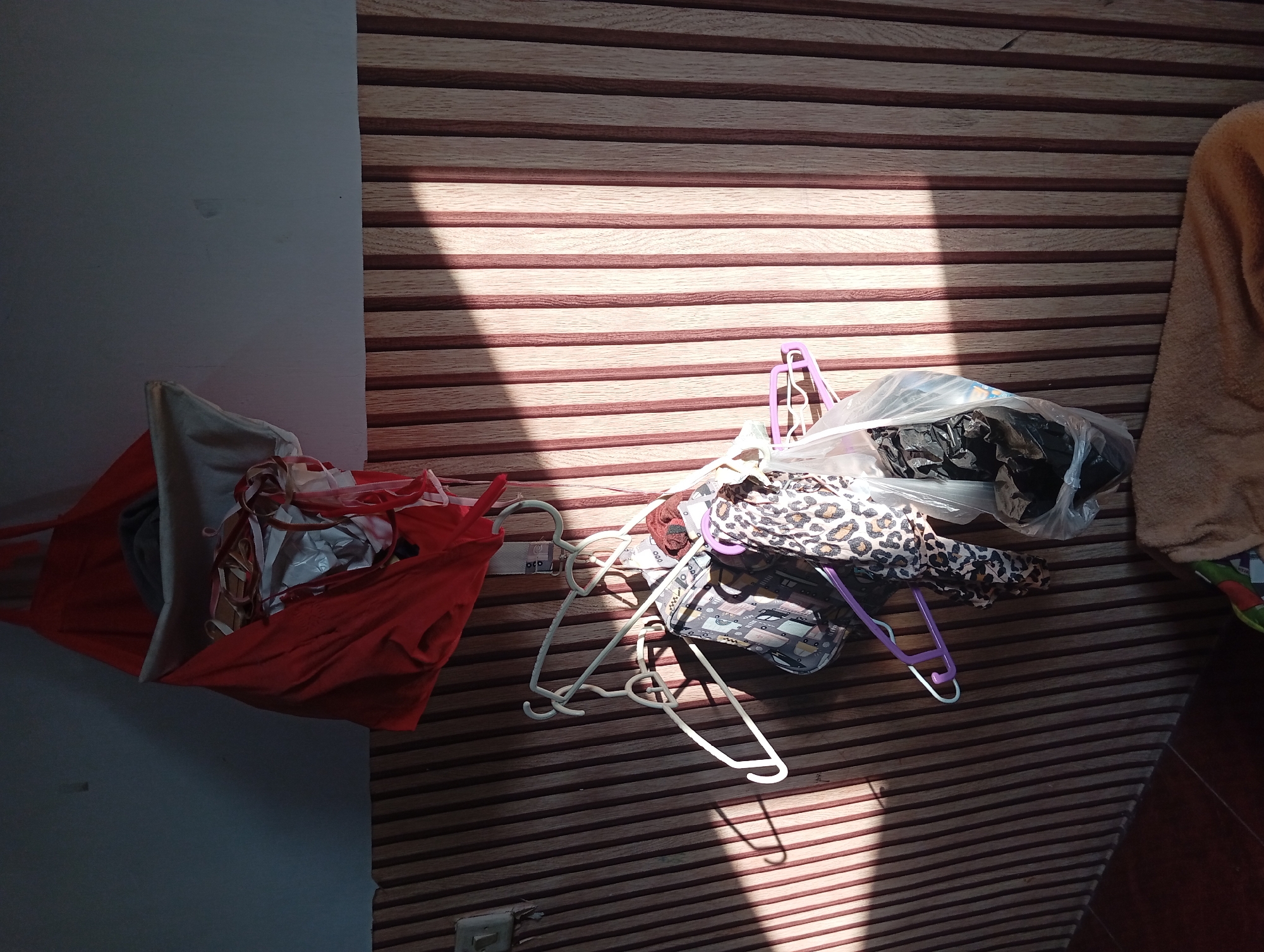 The image depicts an indoor scene, likely a laundry area or a storage space. A red fabric bag, possibly a laundry bag, is hung from the wall near the ceiling. Below it, several clothes hangers are suspended from a rack. Various items of clothing are hanging on the hangers, including patterned fabrics like leopard print and a design with buildings. Some clothes are also contained within a clear plastic bag. The wall is decorated with vertical wooden panels, and a strong beam of sunlight casts diagonal shadows across the scene. The lower part of the image shows a glimpse of a brown textile, possibly a blanket or a mat, on the floor. The environment appears somewhat cluttered. No people are visible in the image. The location is identified as Jogosari, Indonesia.