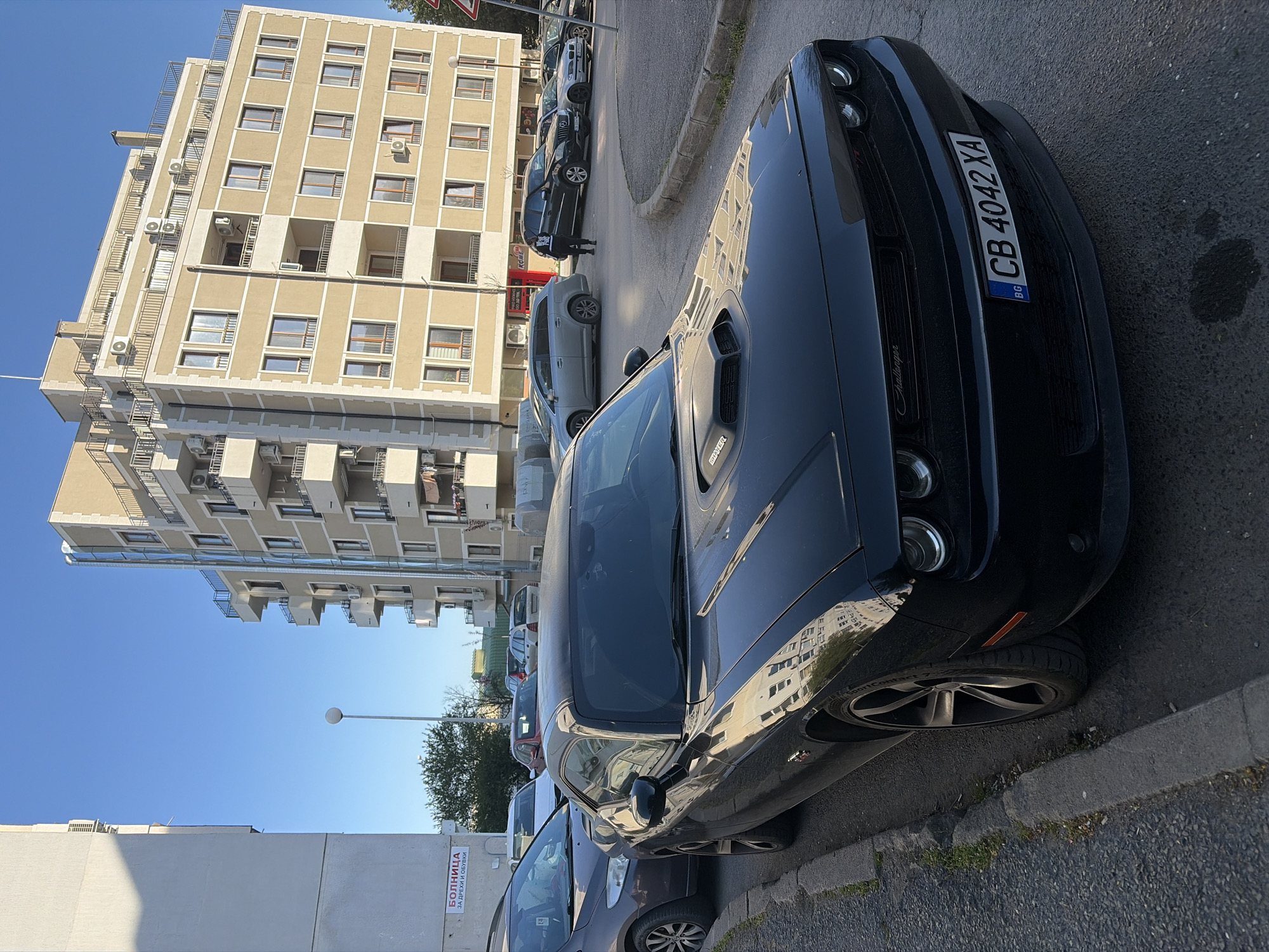 The image shows a black Dodge Challenger parked on a street in Sofia, Bulgaria. The license plate reads "CB 4042 XA." Several other parked cars are visible along the street. A large apartment building is in the background. The sky is blue, suggesting it is daytime. There is a street sign visible on the building on the left. The image suggests a typical urban scene.