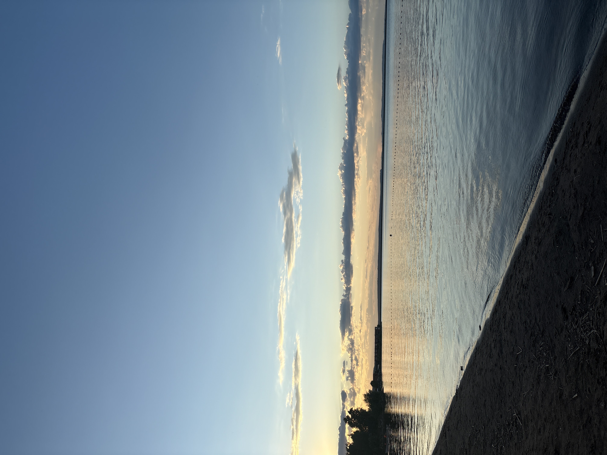 This tranquil landscape captures a wide expanse of water under a serene sky, likely at sunrise or sunset. The sky, occupying the upper portion of the frame, transitions from a deep clear blue overhead to soft golden and orange hues near the horizon, where scattered, elongated clouds catch the warm light. A distinct horizon line separates the sky from the calm water, with a faint, dark distant shoreline or landmass visible along it.
The large body of water, possibly a lake or a wide river in Chaikovsky, Russia, is largely still with gentle ripples, beautifully reflecting the sky's colors, especially the golden glow from the low sun. A prominent detail on the water is a long, dotted line of small, dark objects, likely buoys or markers, stretching horizontally across the surface, indicating a boundary or path. On the distant left horizon, a dark silhouette of trees or a structure is visible, mirrored faintly in the water.
In the foreground, a dark, sandy or earthy beach or shoreline slants diagonally from the lower right, meeting the water's edge. This dark ground contrasts sharply with the illuminated water and sky. No people, animals, or visible text are present, and no specific activity is taking place, contributing to the peaceful and contemplative atmosphere of the scene. The warm, soft lighting underscores the serene mood of the early morning or late evening.