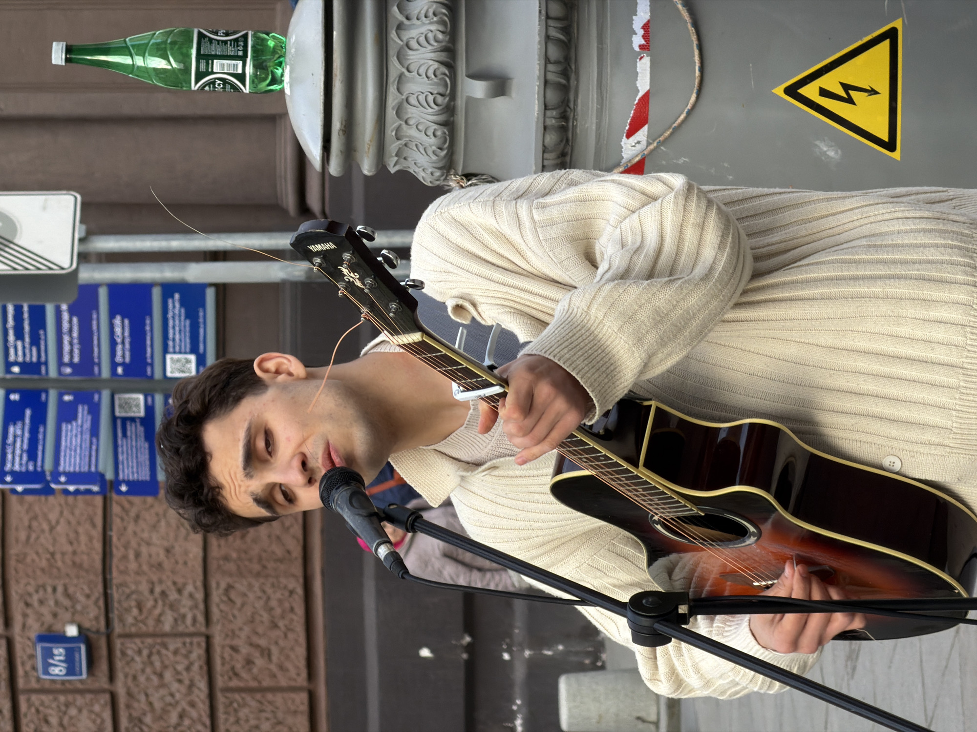 A man plays a guitar and sings into a microphone in Moscow, Russia. The man has dark hair and is wearing a beige sweater. He is standing next to a lamppost with a yellow and black electrical hazard sign. A green bottle of water is sitting on top of the lamppost. A set of blue and white signs can be seen in the background. The scene appears to be outdoors on a city street.
