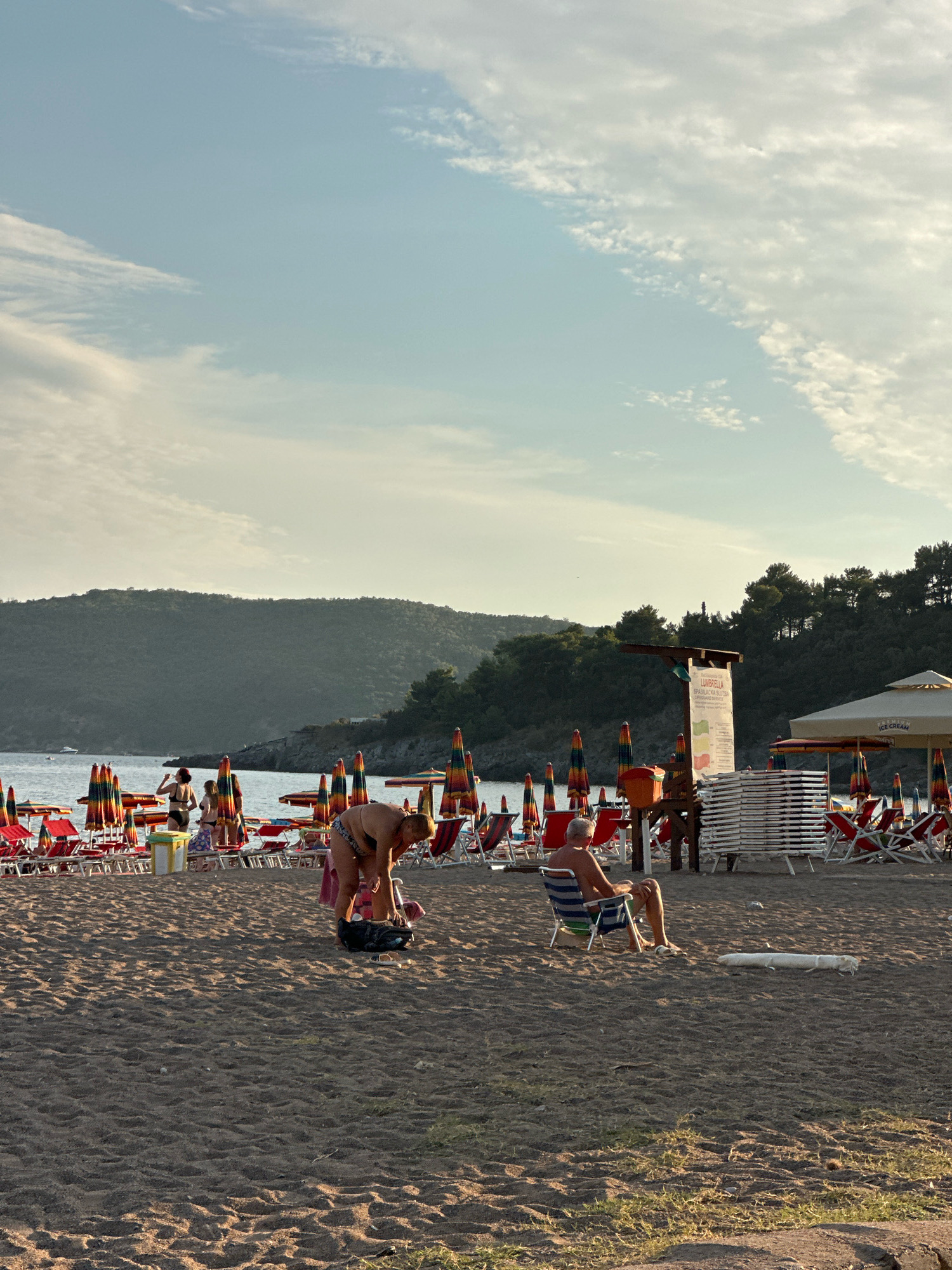 The image depicts a sun-drenched beach scene in Boreti, Montenegro. In the foreground, the sandy beach is marked by the shadows of beach umbrellas, and a few people are engaged in leisure activities. A woman in a bikini bends down to pick up a bag, while a man sits in a striped beach chair, facing away from the camera. Further back, more beach chairs and umbrellas are arranged in neat rows, suggesting a popular tourist spot. The background features the calm blue sea, with a distant green, tree-covered hill rising behind it. The sky is partly cloudy, with a gentle gradient of blue and white. The overall atmosphere is one of relaxation and summer vacation.