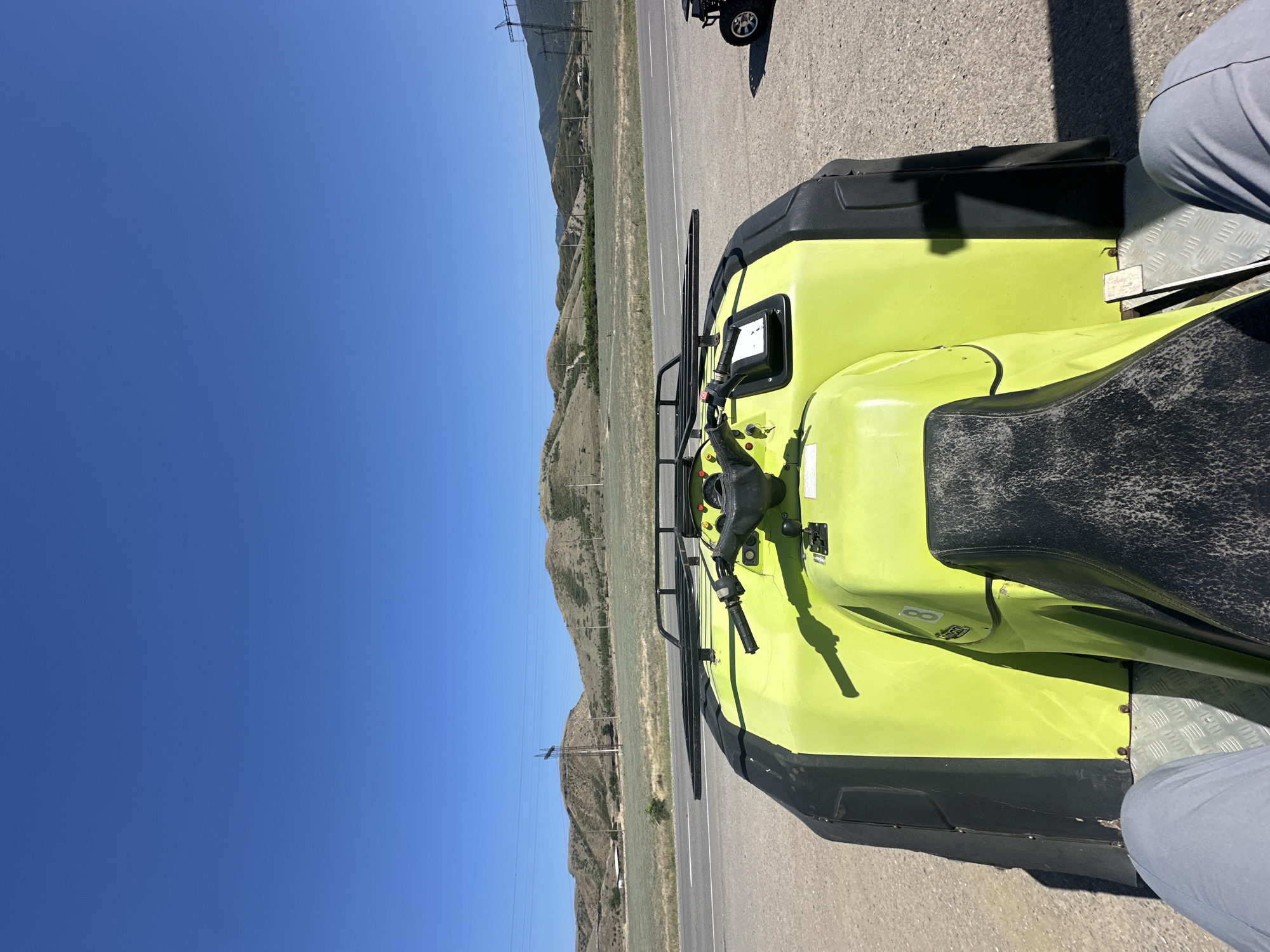 The image captures a first-person perspective from an all-terrain vehicle (ATV) on a clear, sunny day. The ATV, predominantly lime green with black accents, is parked on a gravel surface next to a paved road. The handlebars and a portion of the seat are visible in the foreground. Beyond the ATV, a road stretches into the distance, bordered by a grassy field and rolling, arid hills. Power lines and utility poles dot the landscape, with a few distant structures suggesting a rural or semi-rural environment. The sky is a vibrant, cloudless blue. Another ATV is partially visible to the right. The location is identified as Miatli, Russia.