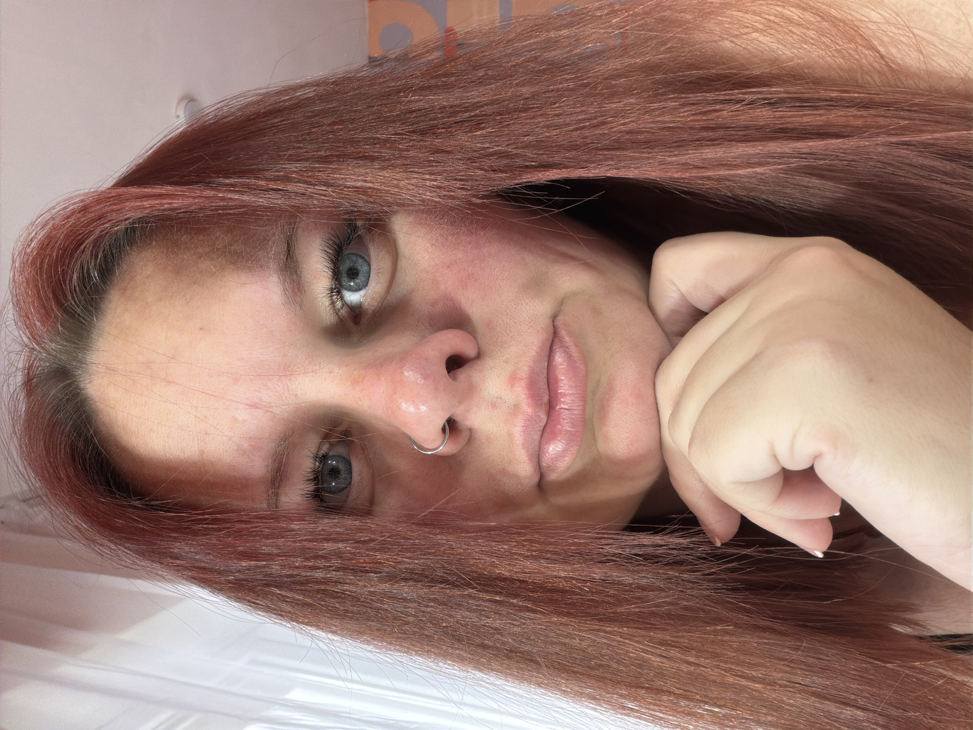 A close-up, rotated portrait captures a woman with reddish-brown hair and light blue eyes. She has a nose ring and her right hand is positioned near her cheek. Her skin tone appears light, with some redness visible on her face. The background is indeterminate, showing a hint of a wall. No information is available in the image to determine any association with the city of Chaikovsky, Russia.