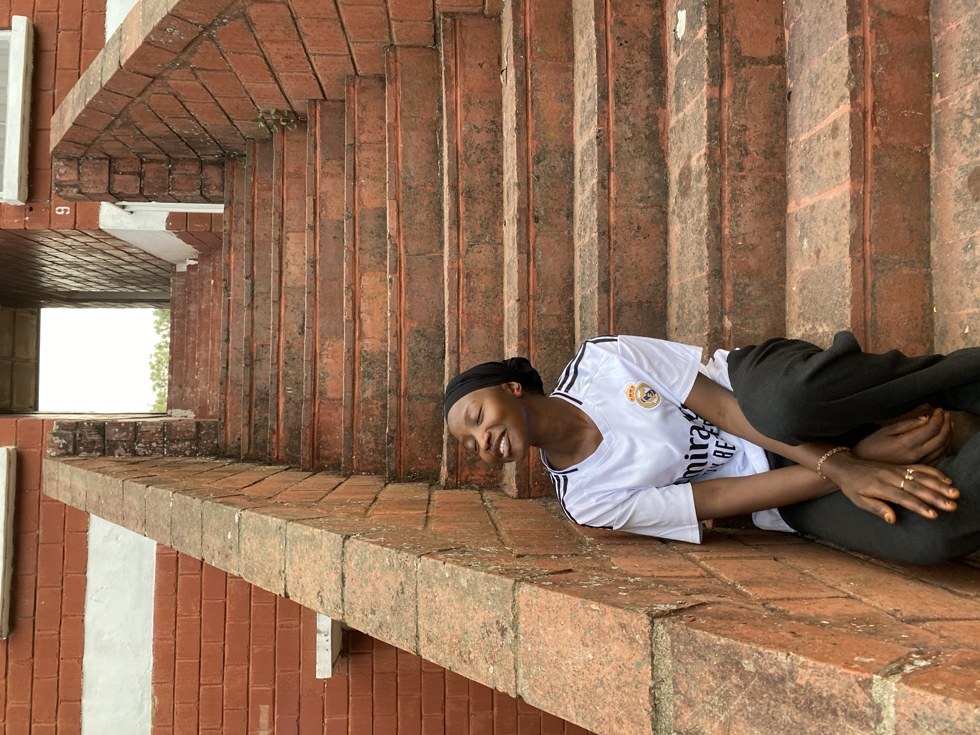 A young Black person is captured in a joyous moment, lying or resting on a weathered brick ledge, smiling widely with closed eyes. They are wearing a white long-sleeved t-shirt with the Real Madrid C.F. logo and "Emirates FLY BETTER" text, black pants, and a black head covering. A gold chain bracelet is visible on their left wrist, and a gold ring is on their right ring finger.
The scene is dominated by reddish-brown brick architecture, likely an outdoor setting in Vom, Nigeria. Behind the person, a series of brick steps or vertical sections ascend, showing signs of wear, dirt, and possibly moss. To the left, a white window frame is partially visible against a brick wall. A small green plant grows out of a crack in the brickwork near the top. The image is taken during the daytime, with soft, even lighting, suggesting an overcast sky or a well-shaded area. A number, appearing as "96" when viewed upright, is stenciled in white on a brick column in the upper left background. The overall impression is one of candid happiness within a rustic, aged architectural environment.
