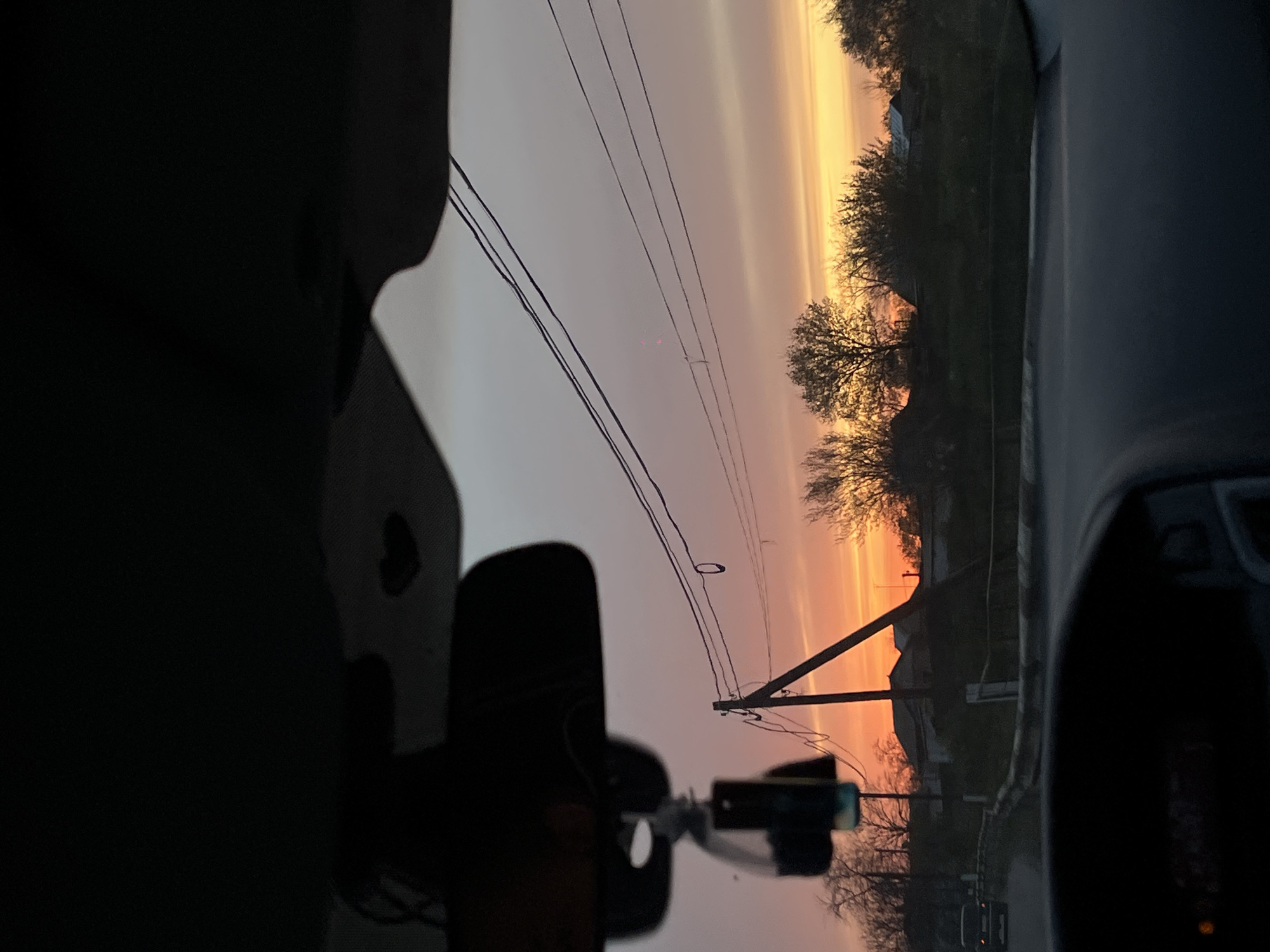 This image captures a view from inside a vehicle, looking out at a sunrise or sunset over a landscape in Alexandrovka, Russia. The sky is a prominent feature, displaying a vibrant gradient of colors, from bright orange and yellow near the horizon on the right, transitioning to soft pinks and grays across the rest of the sky. Long, horizontal cloud streaks are visible in the upper sky, catching the light.
In the midground, several bare trees are silhouetted against the colorful sky, suggesting late autumn, winter, or early spring. Utility poles with multiple power lines stretch across the scene, adding an element of human infrastructure to the natural beauty. Faint outlines of distant buildings, possibly houses, can be seen nestled among the trees, indicating a rural or semi-rural setting.
The foreground is dominated by the dark interior of the car, including parts of the dashboard and the silhouette of a rearview mirror on the left. A smaller device, possibly a dashcam or phone mount, is also silhouetted towards the bottom-left. The overall atmosphere conveys a sense of peaceful observation during the transitional period of dawn or dusk. No visible text is discernible in the image.