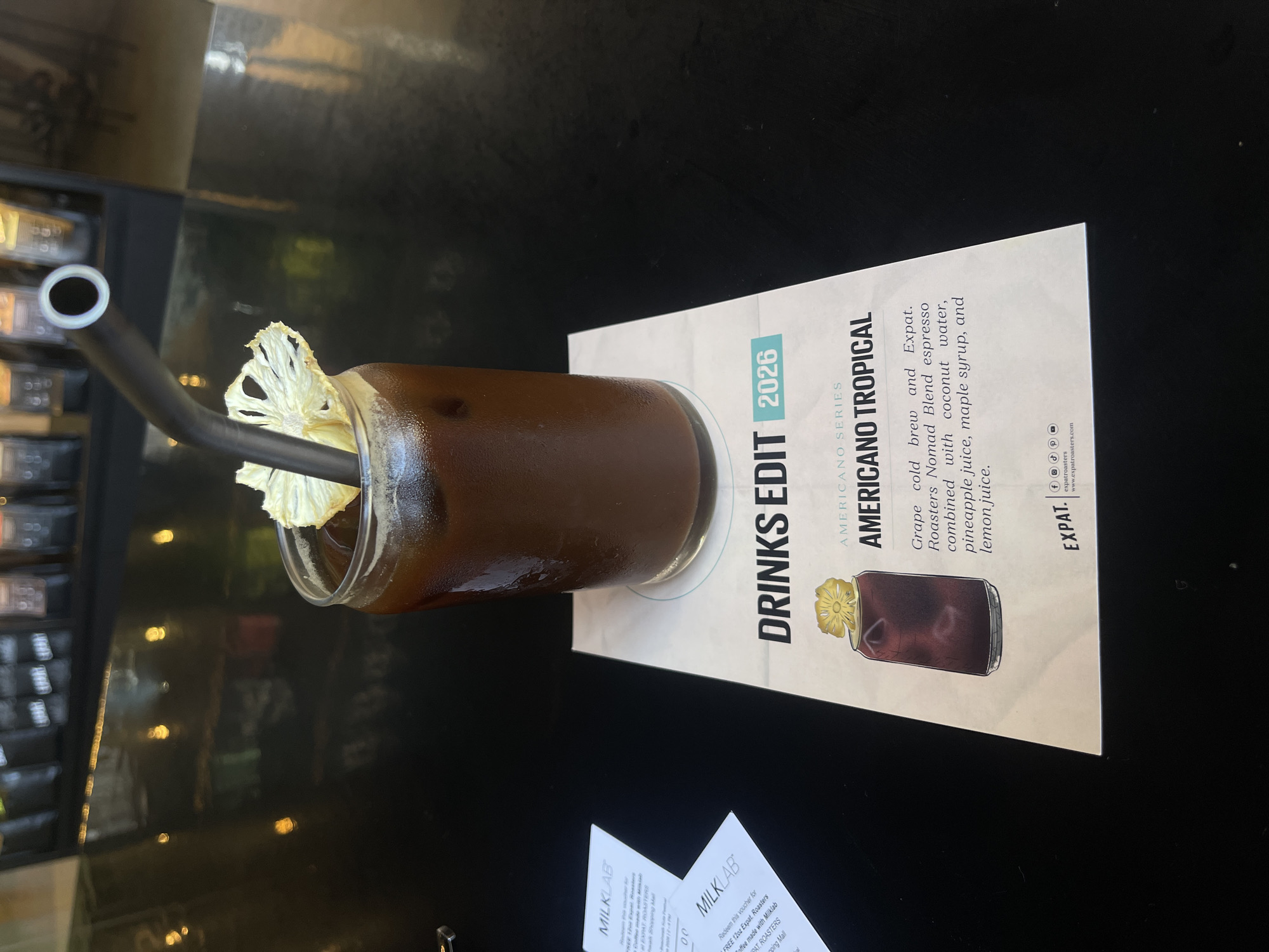 The image captures a static scene featuring a "Americano Tropical" drink on a dark, reflective table, likely within a cafe setting in Kuta, Indonesia. The main subject is a clear glass filled with a dark, iced beverage, garnished with a dried pineapple slice on its rim and containing a dark, bent straw.
Beside the drink is a white menu card, prominently displaying "DRINKS EDIT 2026." The card describes the drink as part of the "AMERICANO SERIES" and lists its ingredients: "Grape cold brew and Expat. Roasters Nomad Blend espresso combined with coconut water, pineapple juice, maple syrup, and lemon juice." The card also features a small illustration of the drink and branding for "EXPAT." and "www.expatroasters.com," along with social media icons.
In the bottom left corner, partially visible, are white cards with "MILKLAB" branding. One card mentions a voucher for "FREE 12oz Expat. Roasters coffee made with Milklab" at "EXPAT. ROASTERS" located in a "shopping Mall."
The background is dark and out of focus, showing blurred reflections and what appears to be shelves or displays, consistent with a cafe environment. The scene is well-lit, but the time of day is not discernible. The overall impression is a promotional or still-life photograph showcasing a specialty coffee drink with unique tropical ingredients and clear branding information. No people are visible.