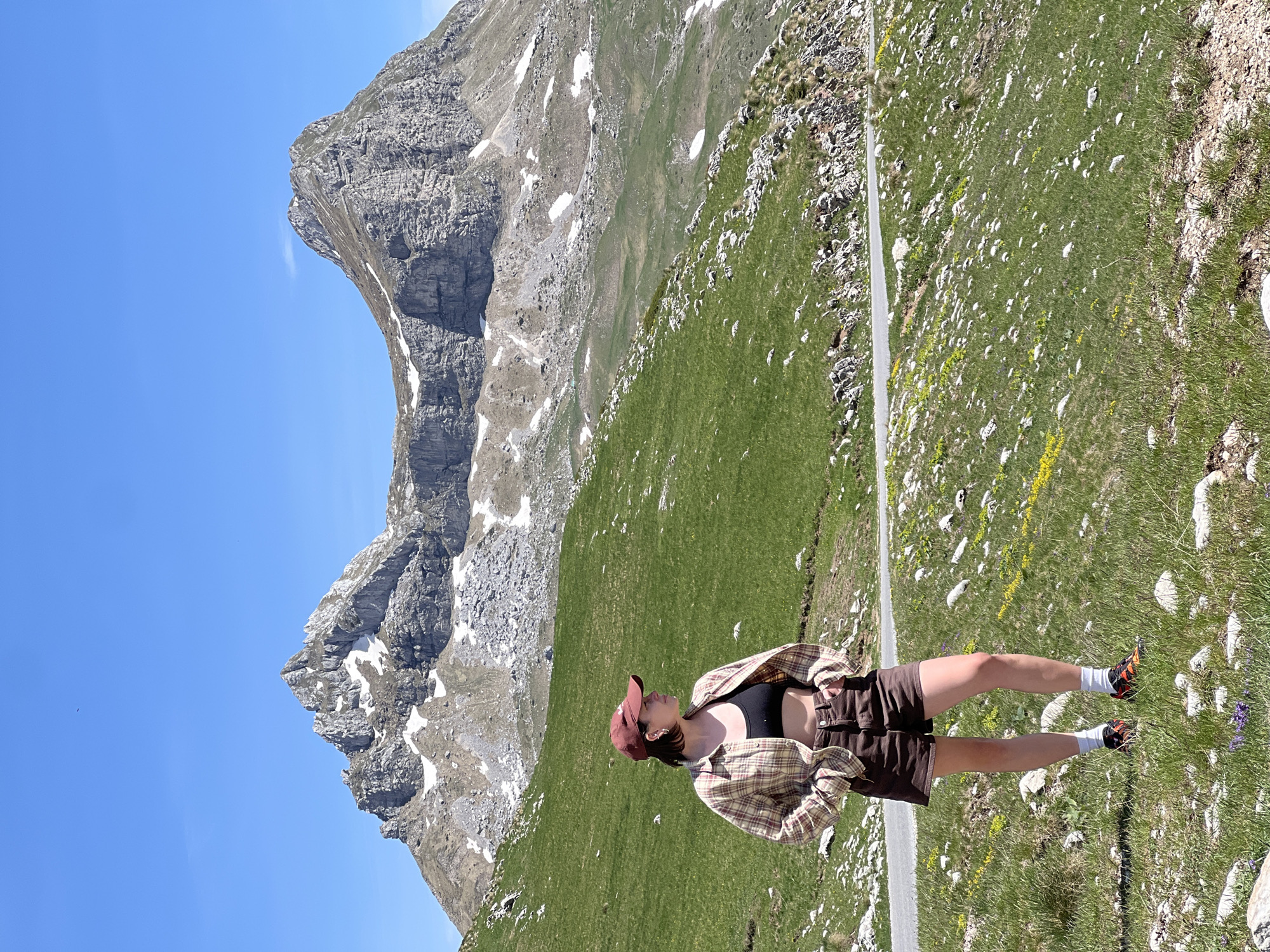The image depicts a woman standing in a mountainous landscape in Dubrovsko, Montenegro. She is wearing a brown sports bra, brown shorts, a tan and brown plaid shirt, a red cap, white socks, and dark-colored hiking shoes. The woman is on a grassy slope alongside a road. Behind her, a mountain rises with patches of snow and exposed rock. The sky is blue and clear. The scene is bathed in sunlight.