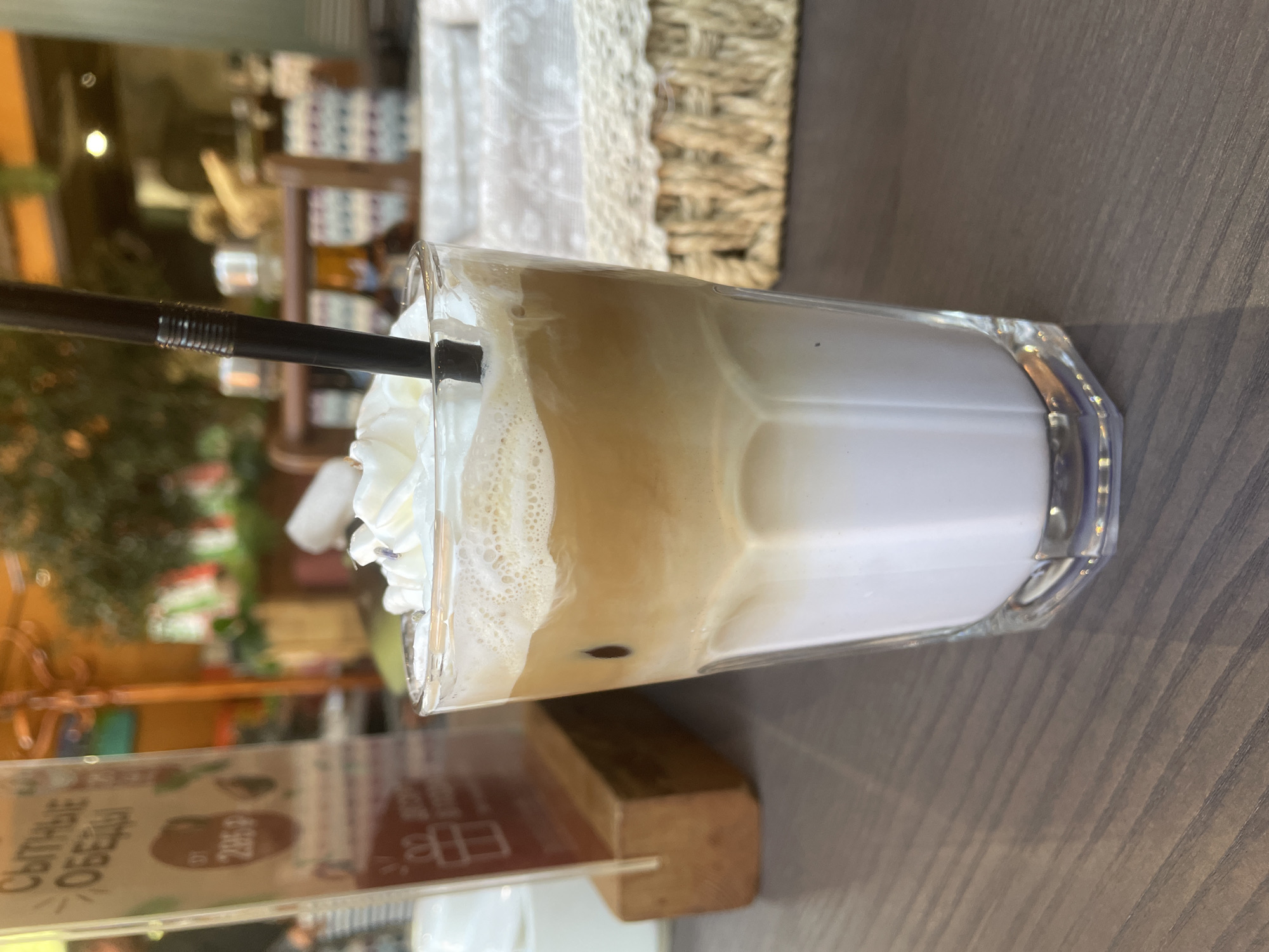 An iced coffee drink topped with whipped cream and a black straw sits on a wooden table in what appears to be an outdoor seating area of a cafe. A menu stand with Russian text and a price of "285 ₽" is visible to the left of the drink. In the background, a wicker basket and greenery suggest a relaxed, possibly European setting. The lighting indicates it is daytime. Without specific landmarks, pinpointing an exact location in Kaliningrad, Russia, is not possible, but the presence of Russian text on the menu is a strong indicator of its geographical context.