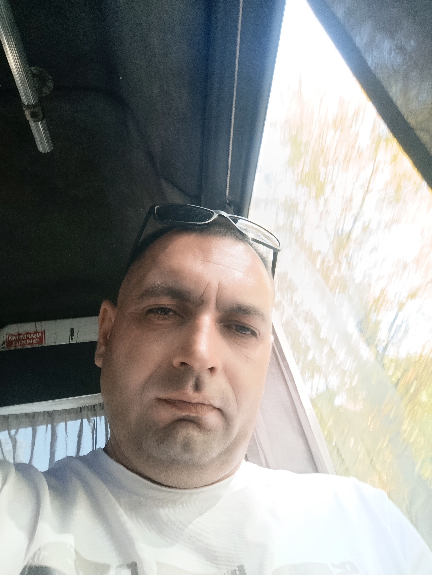 A close-up selfie, taken from a low angle, shows a bald, middle-aged man with dark eyes and a light complexion wearing sunglasses on top of his head and a white t-shirt. He is looking directly at the camera. The background is blurred and appears to be a moving vehicle, possibly a bus or a train, with a window showing trees and sky. A red and white sign with text in what appears to be Cyrillic is visible on the dark interior wall to the left of the man's head. The lighting suggests it is daytime, with bright sunlight filtering through the window. The image is likely taken in Dnipro, Ukraine, based on the presence of Cyrillic text on the sign.