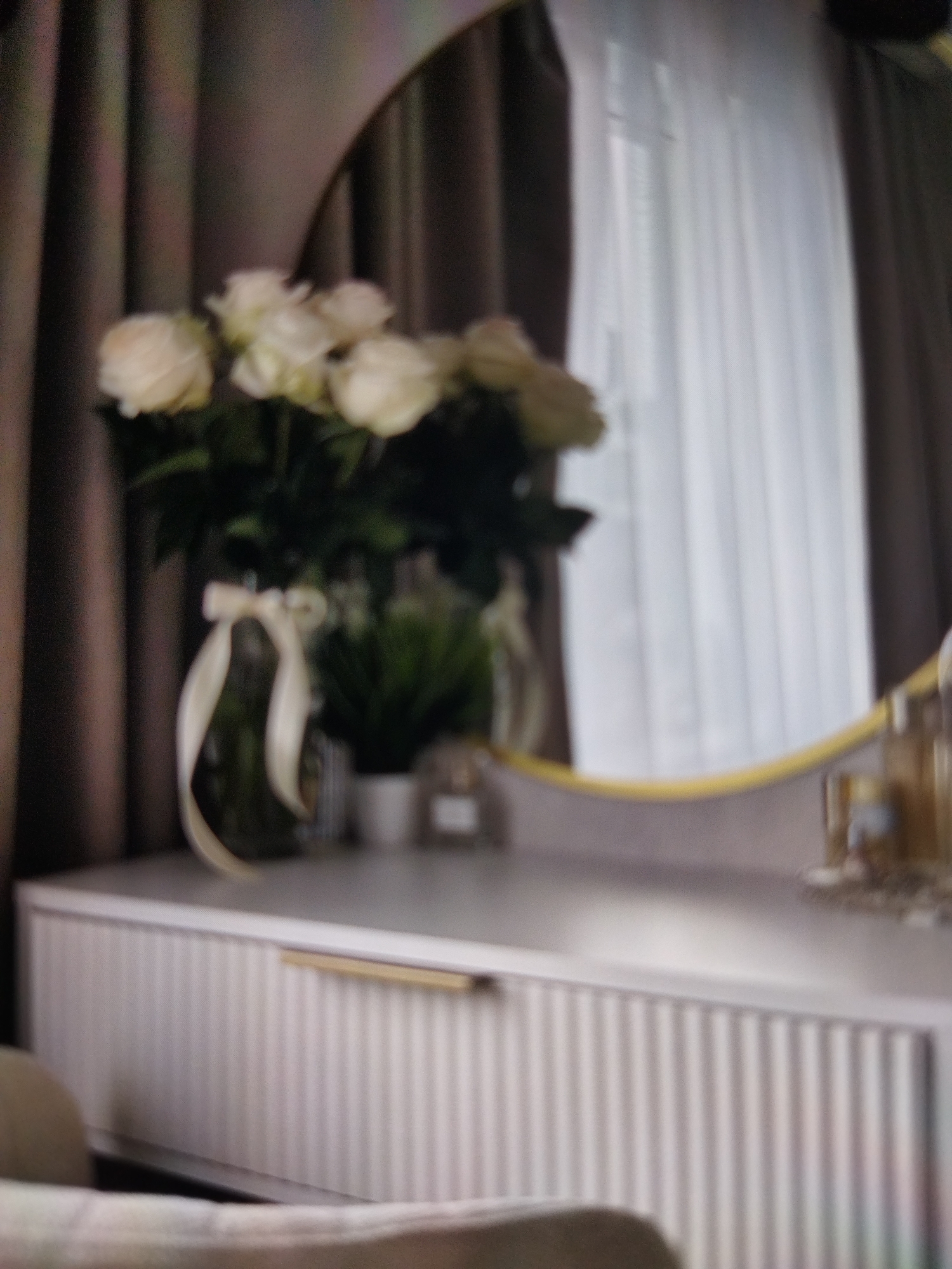 The image depicts a dimly lit indoor scene, possibly a bedroom or dressing area. A bouquet of white roses in a clear glass vase, adorned with a pale ribbon, is placed on a white dresser. Behind the flowers, a large round mirror with a gold rim reflects a sheer white curtain and dark drapes. The dresser itself has a fluted front and a single gold handle. To the right of the mirror, other objects are visible on the dresser, including what appear to be perfume bottles. The overall impression is one of calm and elegance, with soft lighting. The exact time of day is not evident, but the ambient light suggests it could be evening or a cloudy day. The location is Beșalma, Moldova. No people are visible in the image.