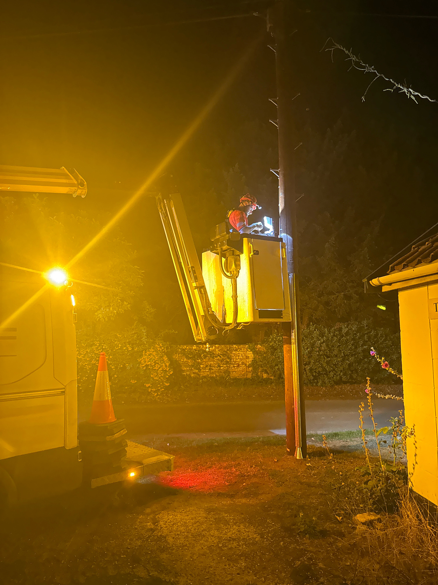 At nighttime in East Suffolk, United Kingdom, a utility worker is performing maintenance on a power line. The worker is situated in the basket of a cherry picker, which is positioned next to a wooden utility pole. The worker is wearing a hard hat and a safety vest and appears to be using a laptop. The scene is illuminated by the bright yellow light from the cherry picker's work lights and a vehicle's headlights. There is a traffic cone on the ground near the vehicle, and a building is visible to the right of the pole. The background consists of trees and foliage, suggesting a rural or semi-rural setting. The ground is covered with gravel and grass.