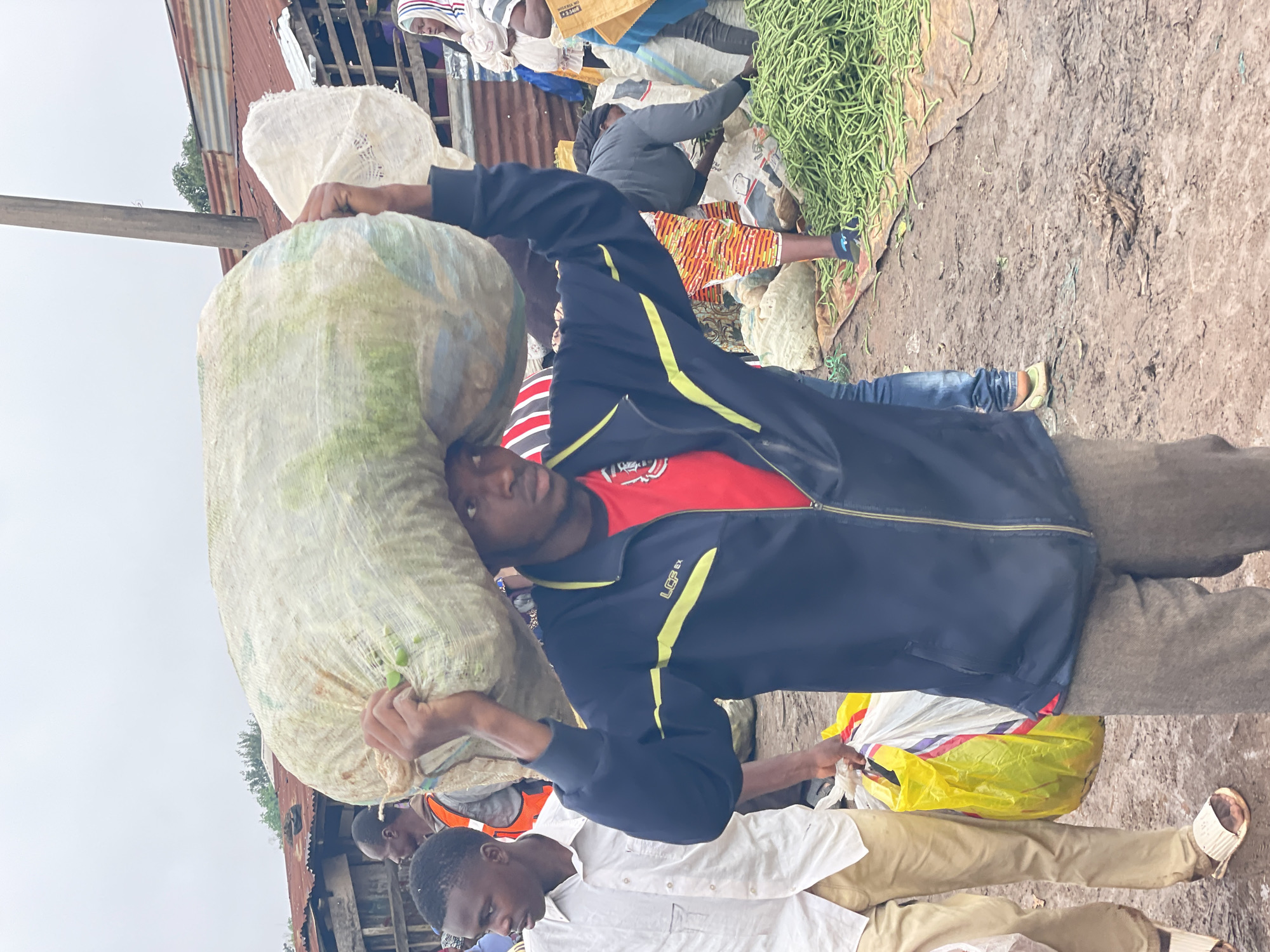 In Gura Top, Nigeria, a man carries a large, light-colored sack of produce on his head through a bustling market. He is wearing a red t-shirt and a dark blue zip-up jacket with yellow accents, and brown trousers. Another man beside him in a white shirt and tan trousers carries a bright yellow bag. In the background, people are seen tending to large piles of green beans. The ground is muddy and littered with debris, indicating a busy, outdoor marketplace. Several market stalls with corrugated iron roofs are visible, suggesting a typical scene of daily commerce.