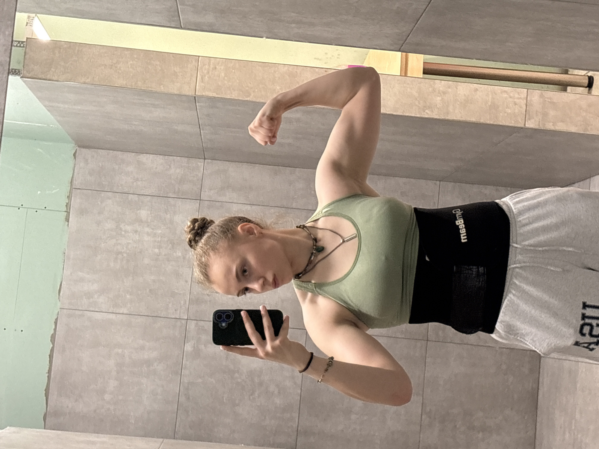 A young woman with fair skin and light brown hair styled in a bun is taking a selfie in what appears to be a bathroom or dressing room with tiled walls. She is wearing a light green tank top, dark colored waist belt, and grey sweatpants with the letters "USA" visible on the right leg. The woman is flexing her right bicep while holding a black smartphone in her left hand. She is wearing several necklaces and bracelets. The environment includes grey tiled walls and what appears to be unfinished drywall on the left side of the frame. A towel rack and a strip of light are above her head. The location is unknown, therefore, I cannot determine if this image was taken in Sofia, Bulgaria.