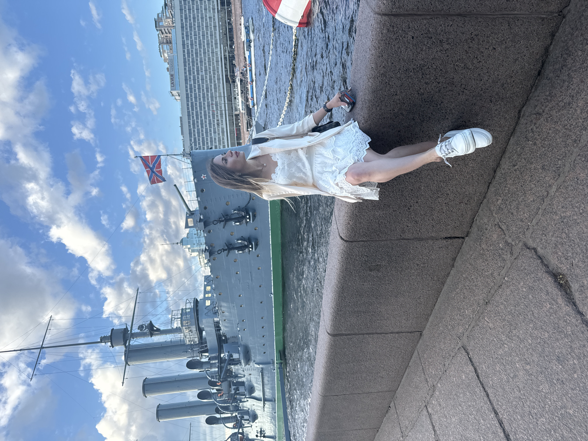 A woman is sitting on a stone embankment overlooking a body of water, with a large, grey, historic ship in the background. The ship has multiple smokestacks and a Russian flag flying from a mast. Behind the ship, a modern building with many windows lines the waterfront. The sky is blue with white clouds. The woman is wearing a white lace dress and a light-colored jacket, and white sneakers. She is looking to her right, away from the camera. The water is choppy, and several smaller boats are visible in the distance. This scene is likely located on the Neva River in Saint Petersburg, Russia, possibly near the Cruiser Aurora, which is a museum ship.