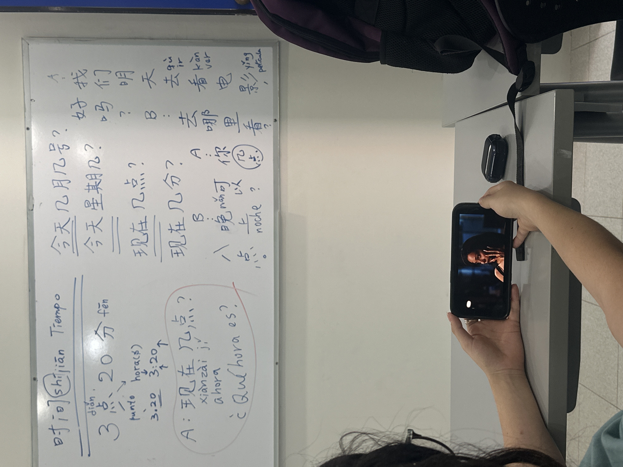 A person is seen from an over-the-shoulder perspective, holding a black smartphone horizontally, seemingly capturing content from a whiteboard. The smartphone's screen displays a close-up image of a person with dark skin and a wide-brimmed hat. Next to the phone on a light grey table, a black earbud case is visible.
The main focus of the image is a whiteboard covered with handwritten Chinese characters, Pinyin, and Spanish translations, indicating a language lesson. Topics include asking and telling time, and making plans. Key phrases visible on the whiteboard are:
* "时间 shíjiān Tiempo" (time)
* "今天几月几号?" (What's the date today?)
* "今天星期几?" (What day of the week is it today?)
* "现在几点?" (What time is it now?)
* "现在几点几分? xiàn zài jǐ, ¿Qué hora es?" (What time and minute is it now?, What time is it?)
* An example of time: "3点20分 fēn" (3:20), with annotations "punto hora(s)", "3.20", "3:20".
* Dialogue related to plans: "好 我们 明天 去 看 电影!" (Okay, let's go watch a movie tomorrow!), "去 哪里 看? ver yíng pelicula" (Where to watch? see movie), "你 几点" (What time you), "晚上 noche" (evening/night).
The setting appears to be a classroom or study environment, likely in San Bernardino Tlaxcalancingo, Mexico, given the context. A dark-colored backpack is partially visible at the top, suggesting a student's presence.