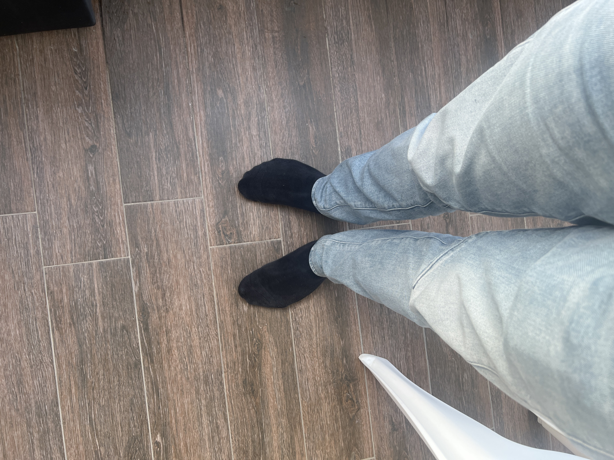 A first-person overhead view depicts a person seated, wearing light blue jeans and black socks, on a wooden-look tiled floor. The lower legs and a portion of a white chair are visible. The location is unknown and there is no evidence to suggest it is in Varna, Bulgaria.