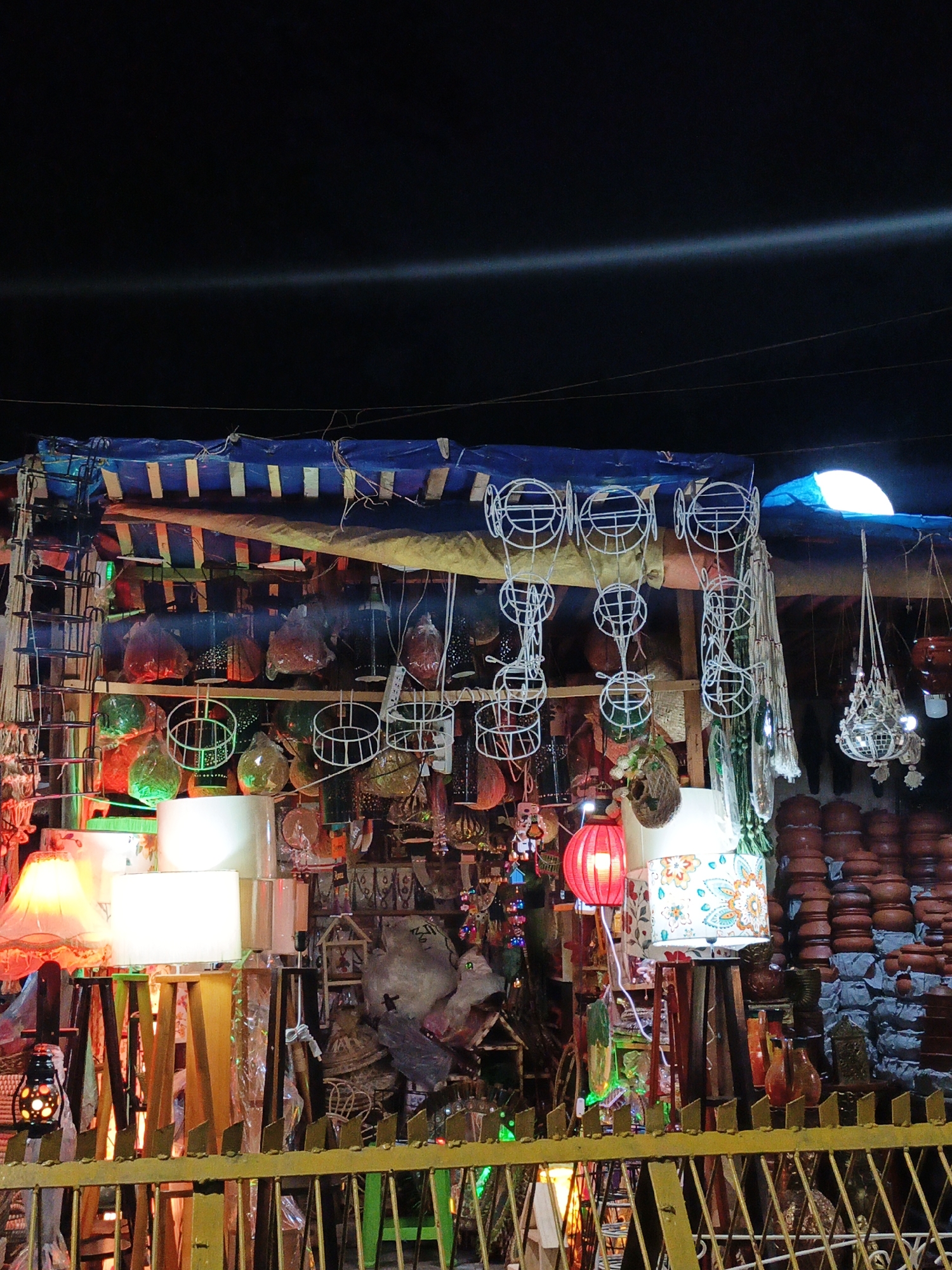 The image captures a vibrant outdoor market stall, likely at night. The stall is brimming with a variety of goods, including lamps of different styles, colors, and sizes, along with decorative items and pottery. The stall is covered by a blue and white striped awning. A yellow metal fence forms the front of the stall. A collection of stacked clay pots sits to the right, and a variety of hanging decorations adorn the stall. Overhead, a bright light source illuminates the scene, with a dark sky in the background.