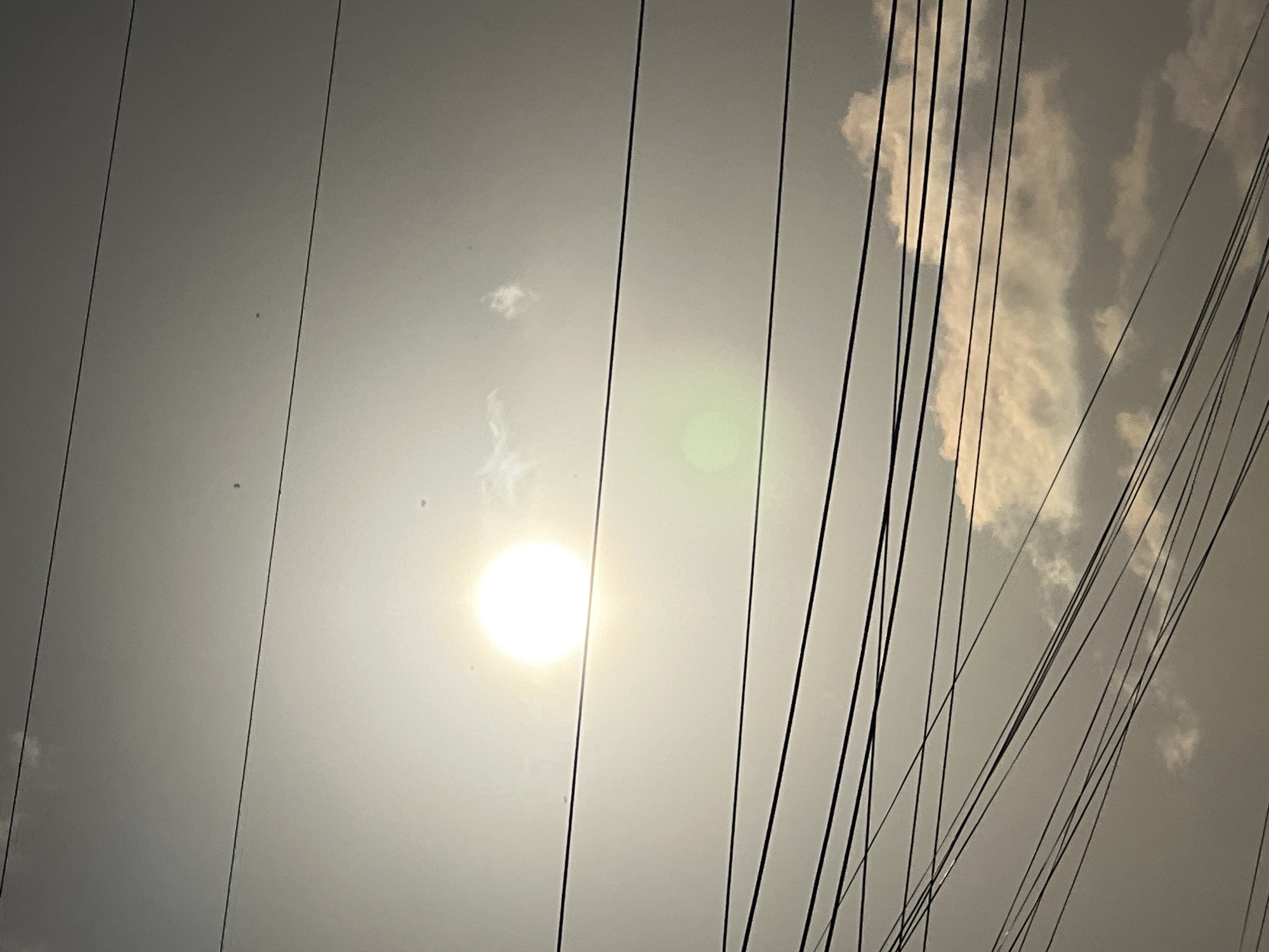 The image depicts a view of the sky above Denpasar, Indonesia, characterized by a bright sun partially obscured by clouds and a dense network of overhead power lines. The sun, a luminous white orb, is positioned in the upper left quadrant of the frame. Several thin, black power lines are diagonally crisscrossed across the image, forming a complex, interwoven pattern against the hazy sky. Below the sun, a soft, green lens flare is visible. In the lower portion of the image, fluffy, light-colored clouds are interspersed with the power lines. The overall lighting suggests it is daytime. There are no people or visible structures in the frame.