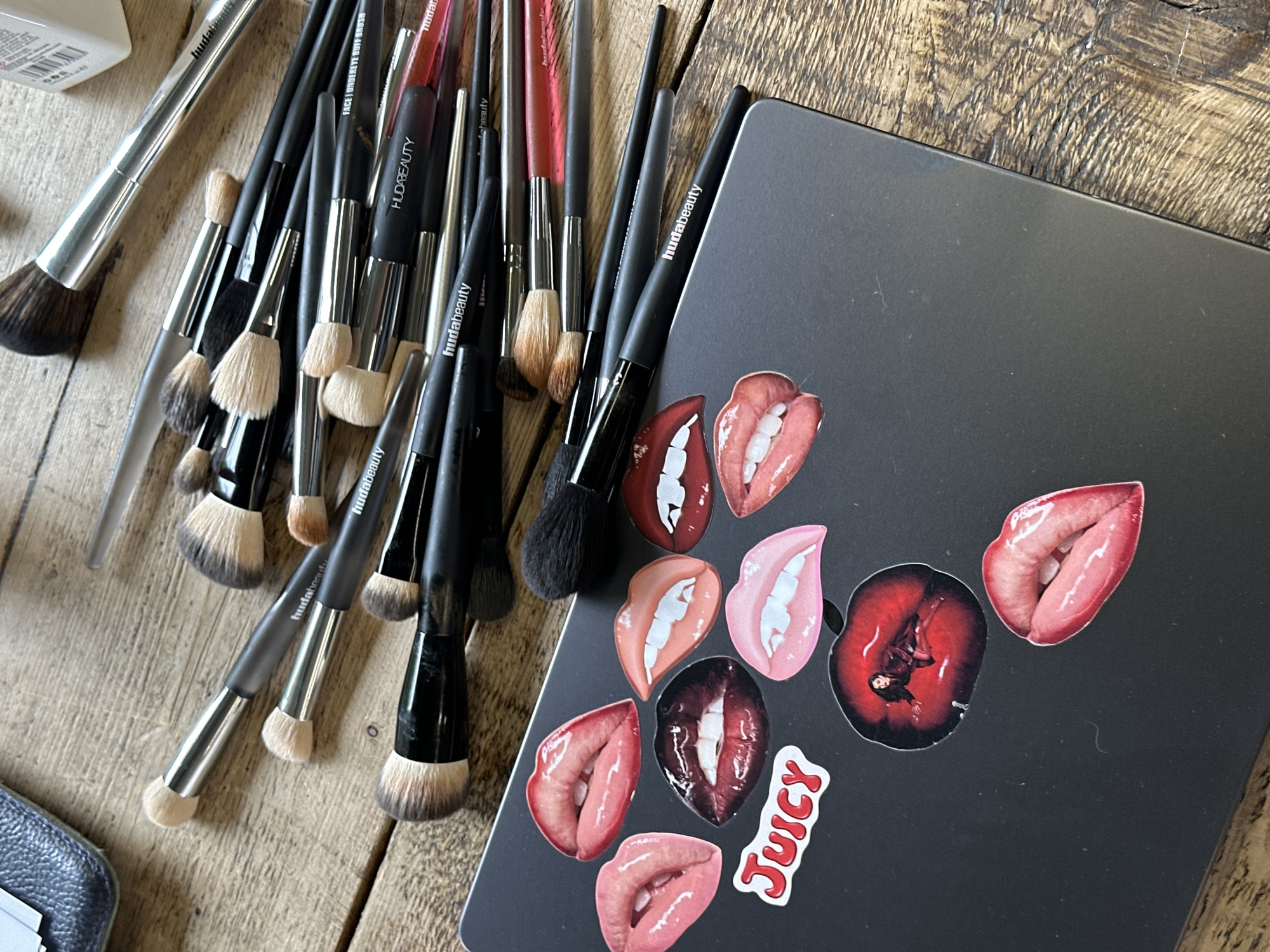 On a wooden surface, likely a table, are a collection of makeup brushes scattered alongside a closed laptop. The brushes are mostly black with silver accents and have "Huda Beauty" printed on them. The laptop is dark gray and adorned with various stickers, mostly featuring depictions of lips in different shades and expressions. One sticker is a red cherry with a figure inside. Another sticker has the word "JUICY" in red letters with a white outline. The scene is casually arranged and appears to be indoors. There is no evidence to suggest this image was taken in London, United Kingdom.